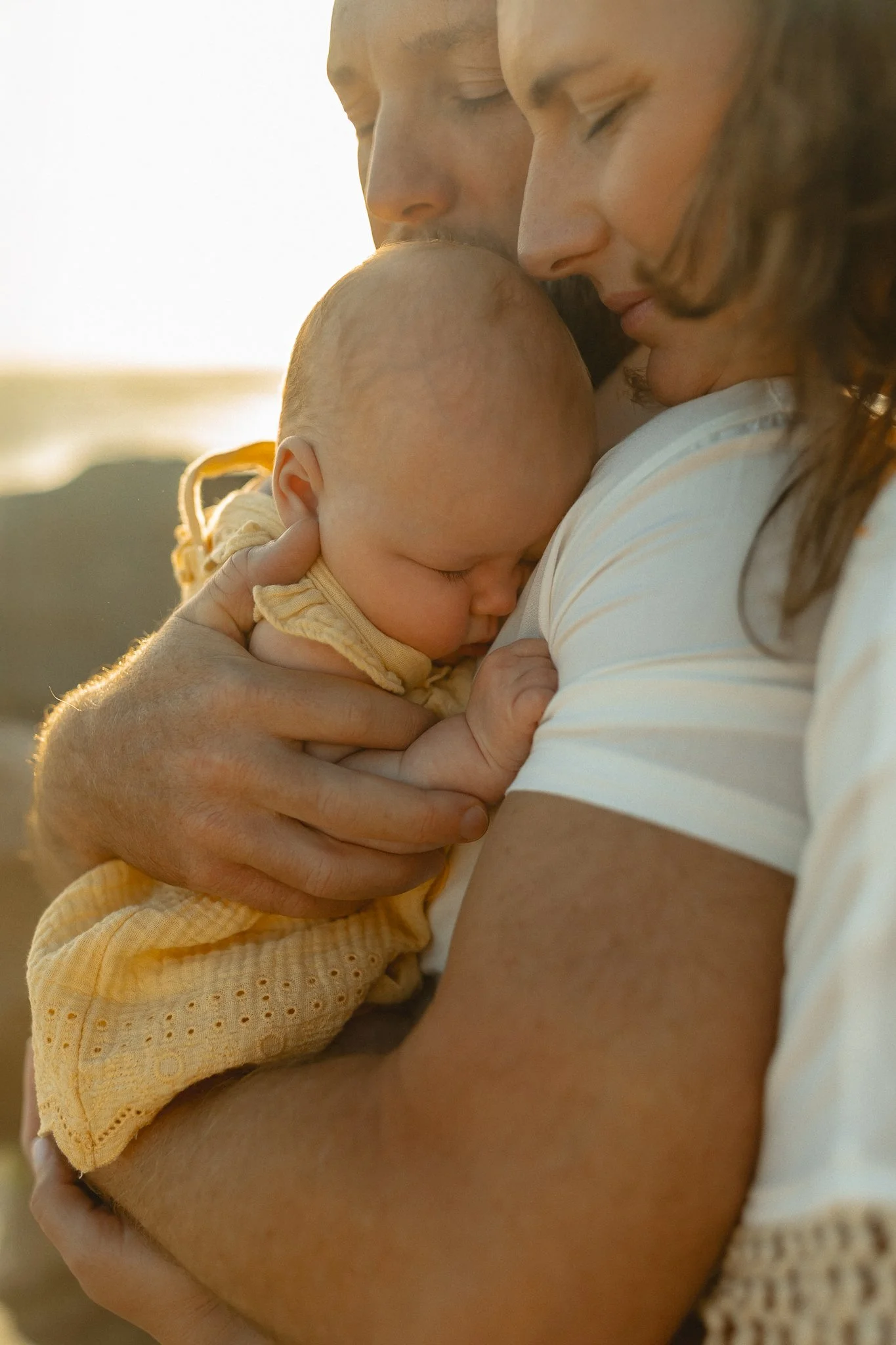 a father holding his newborn baby close to his chest with the mother giving both of them a warm embrace from behind his back