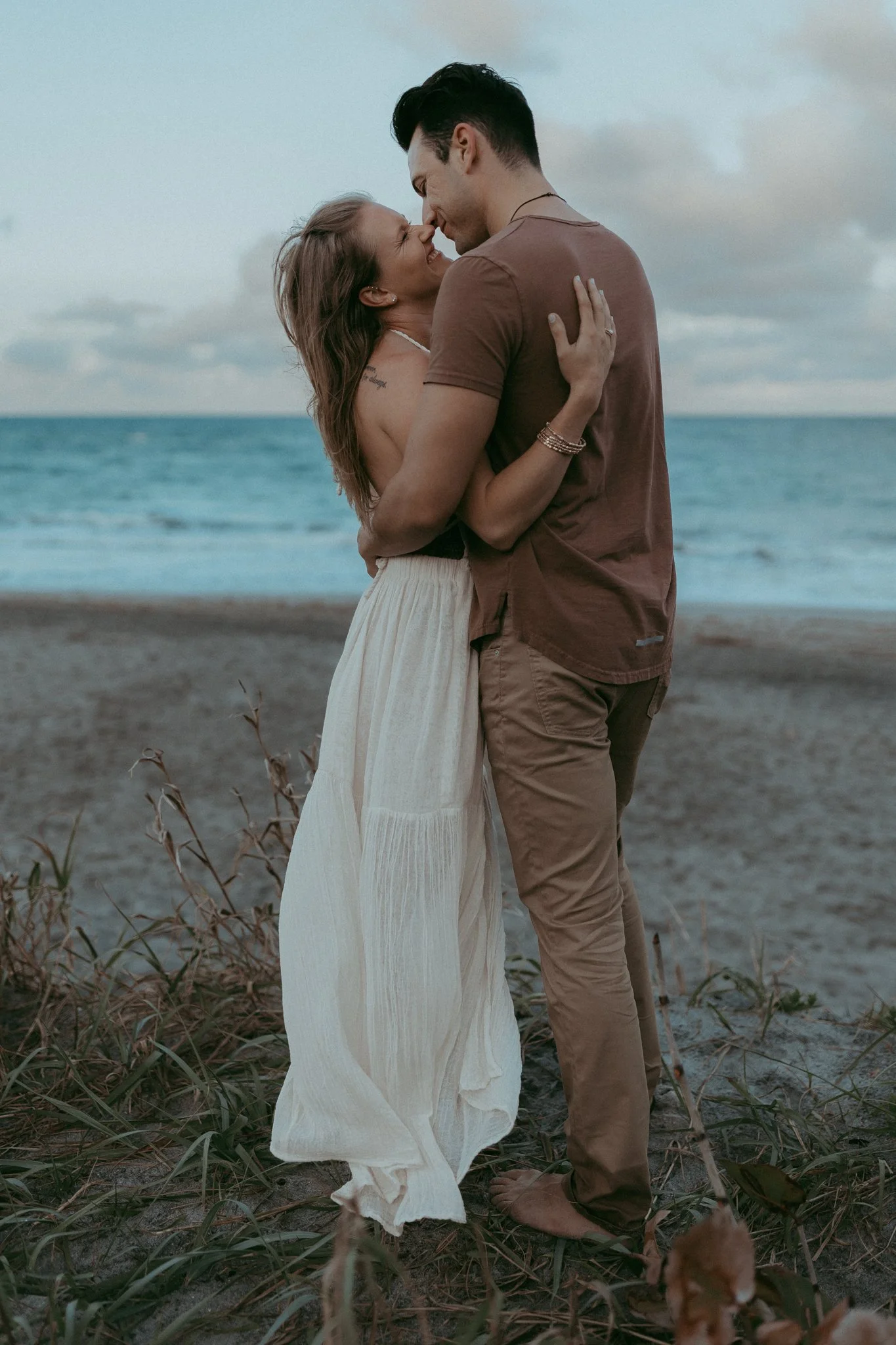 A couple embracing on the beach, smiling and touching foreheads, with the ocean and cloudy sky in the background.