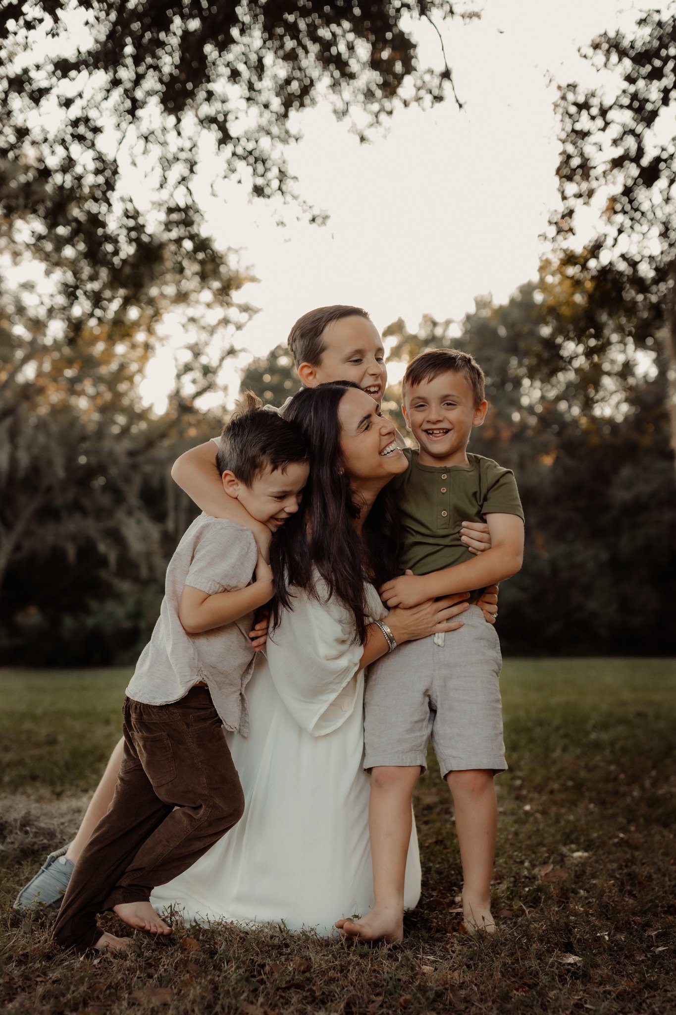 A woman and three young boys laughing and hugging outdoors in a park during the daytime.