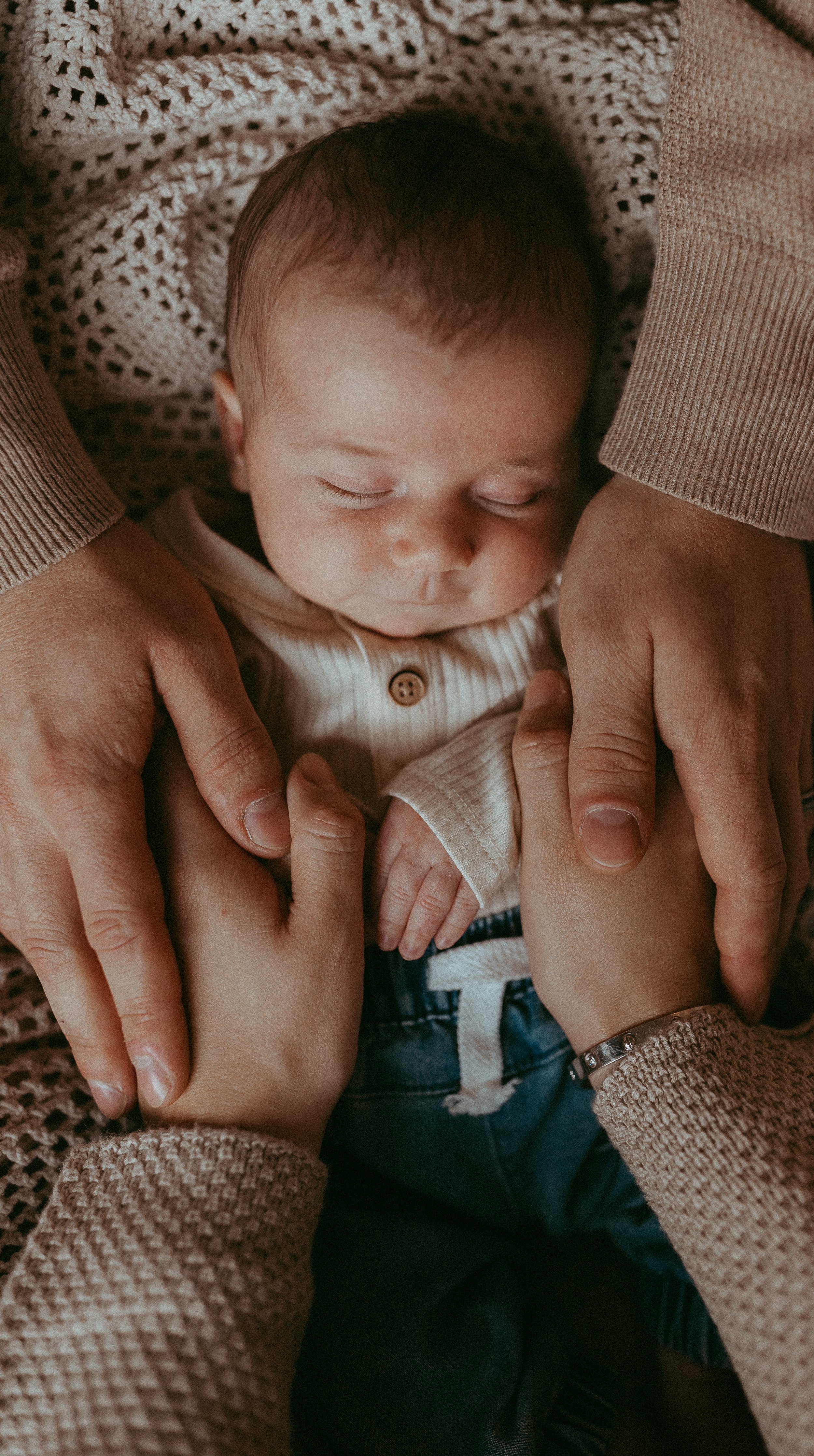A newborn baby sleeping peacefully, surrounded by adult hands gently holding the baby's hands and belly, on a cozy textured blanket.