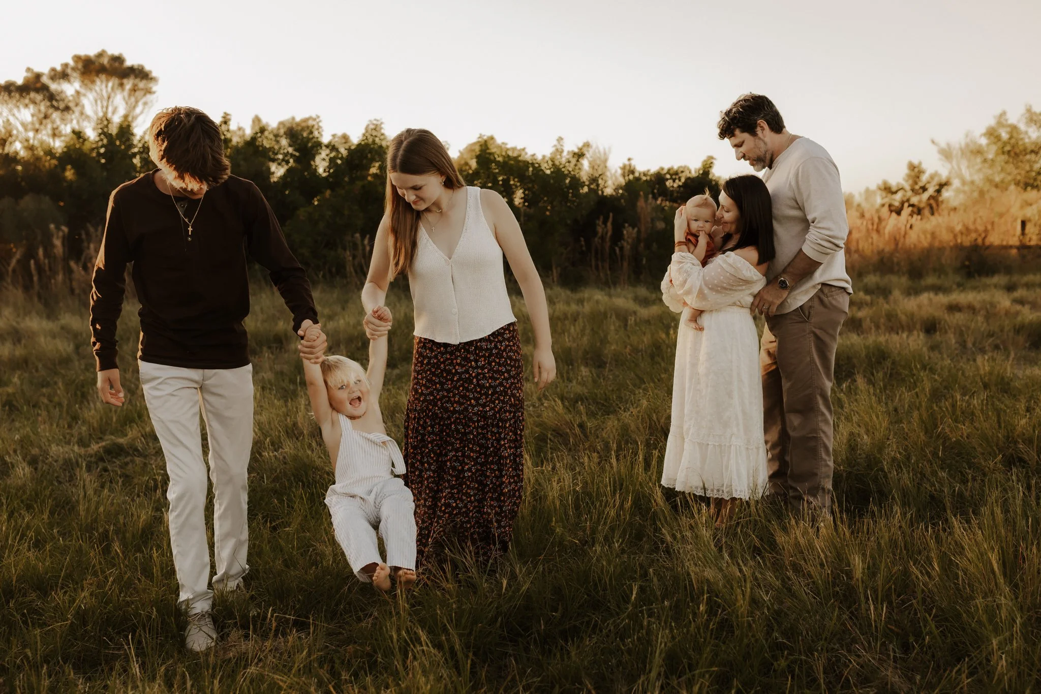 Family of six standing in a grassy field during sunset, holding hands and smiling.