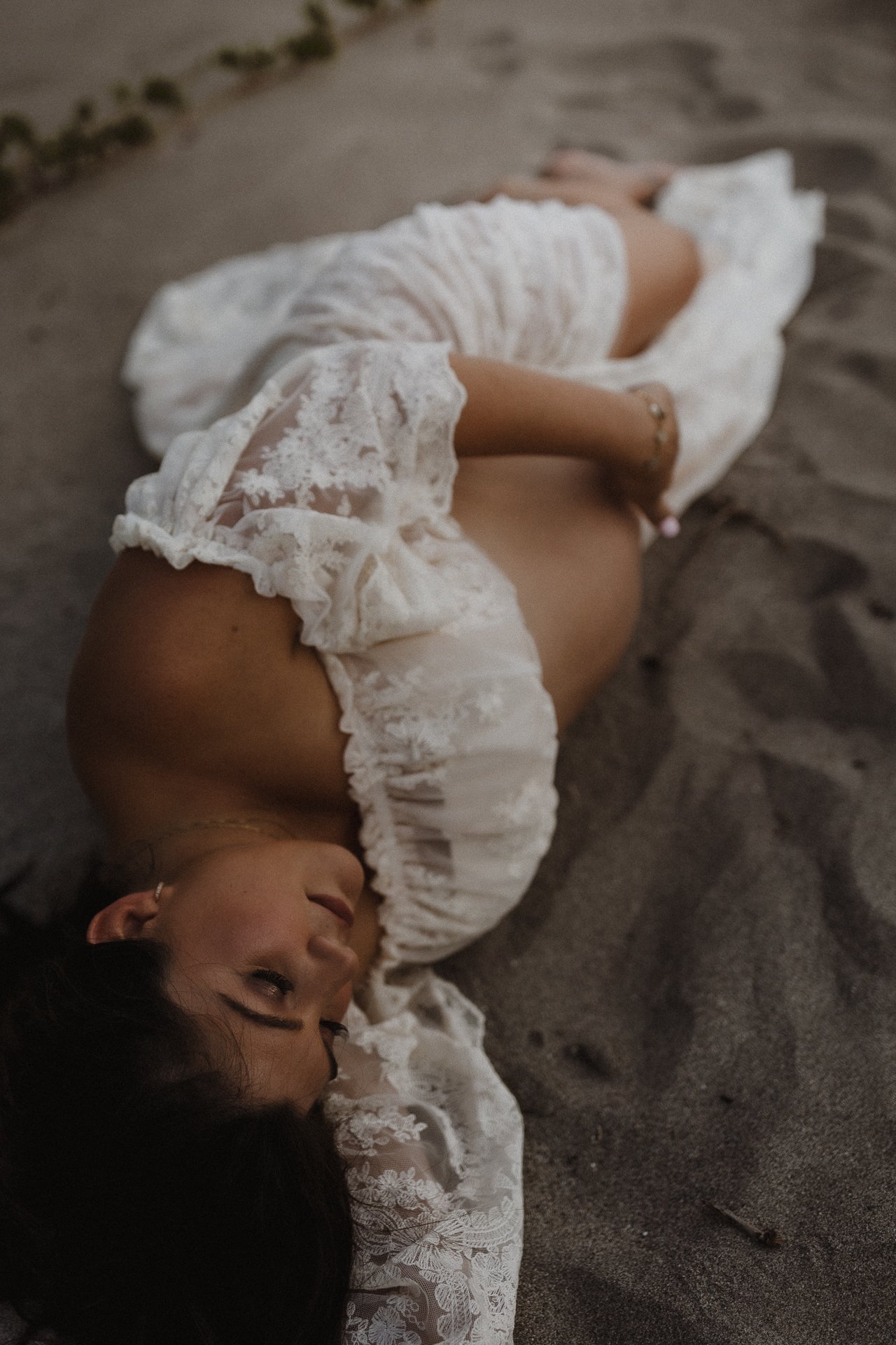 A woman lying on her side on a sandy beach, wearing a white lace dress with puffy sleeves.