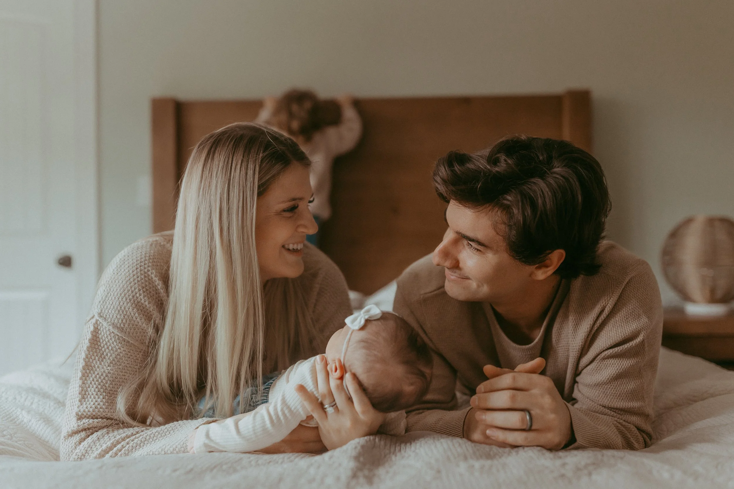 A family of three lying on a bed and smiling at each other, with a baby girl in front of them wearing a white headband.