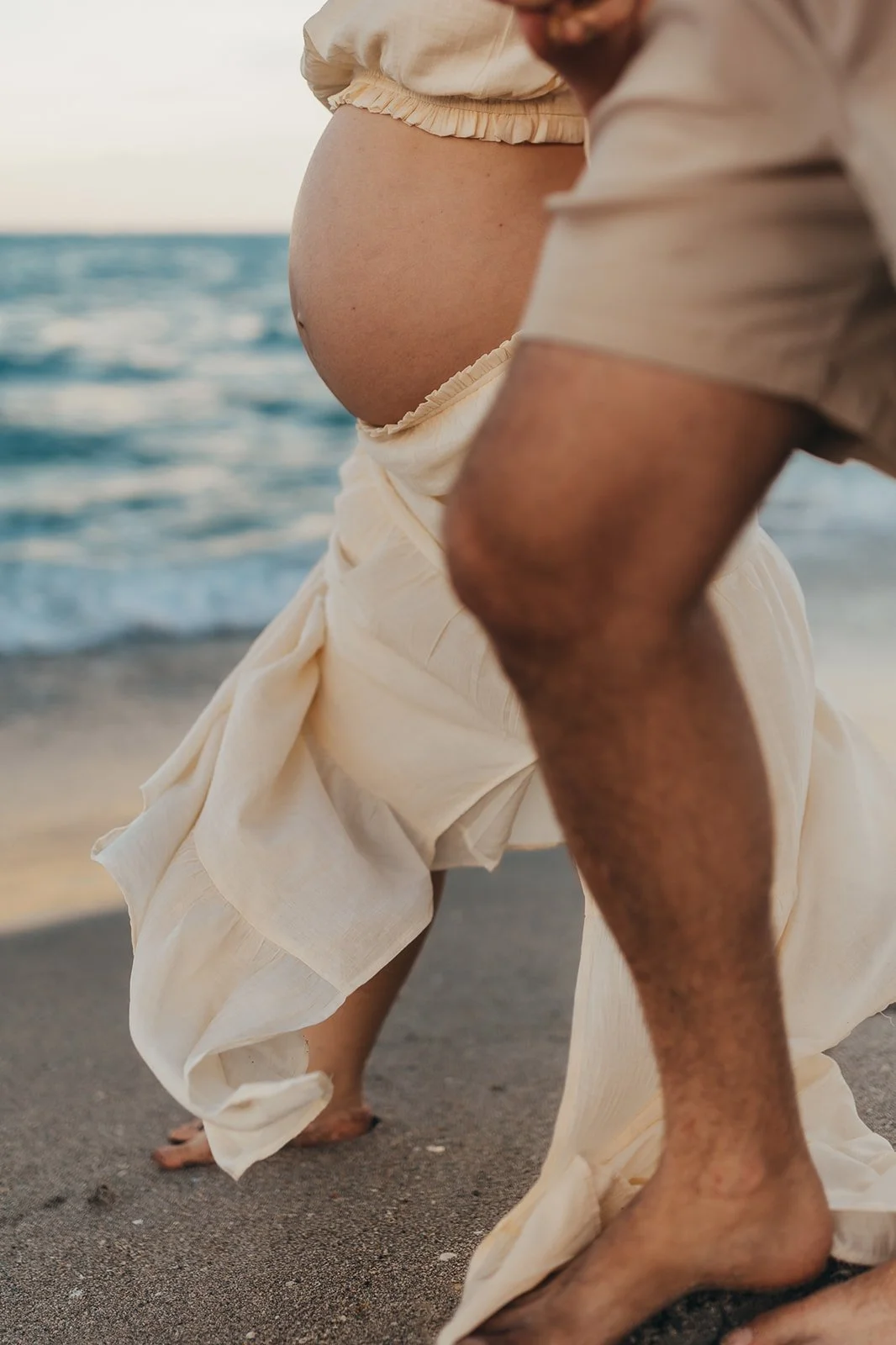 A pregnant woman in a beige dress kneeling on a beach, with a man leaning in close beside her, near the ocean.