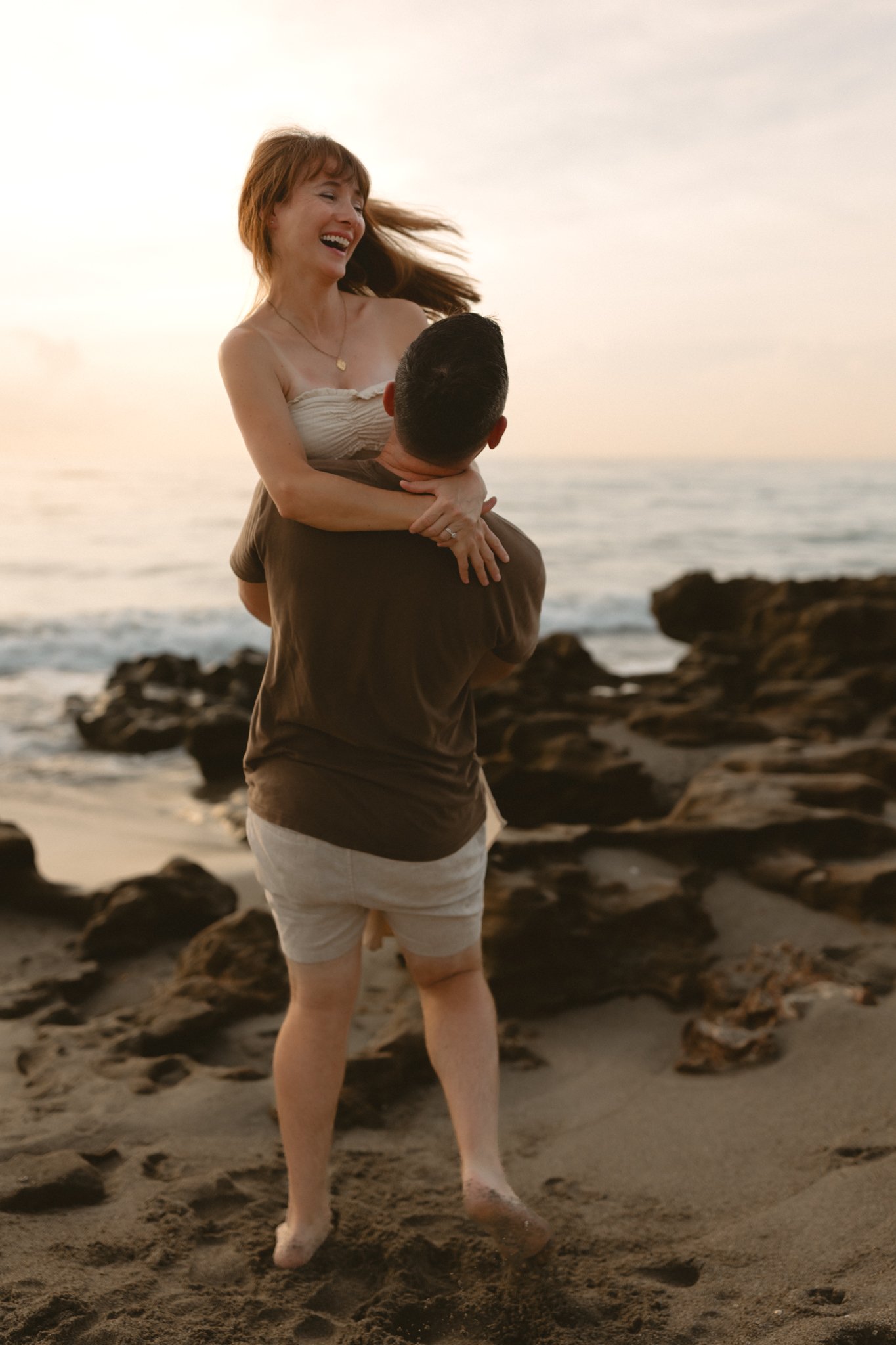 A woman with red hair is being lifted by a man onto his shoulders at the beach, both smiling and enjoying the sunset.
