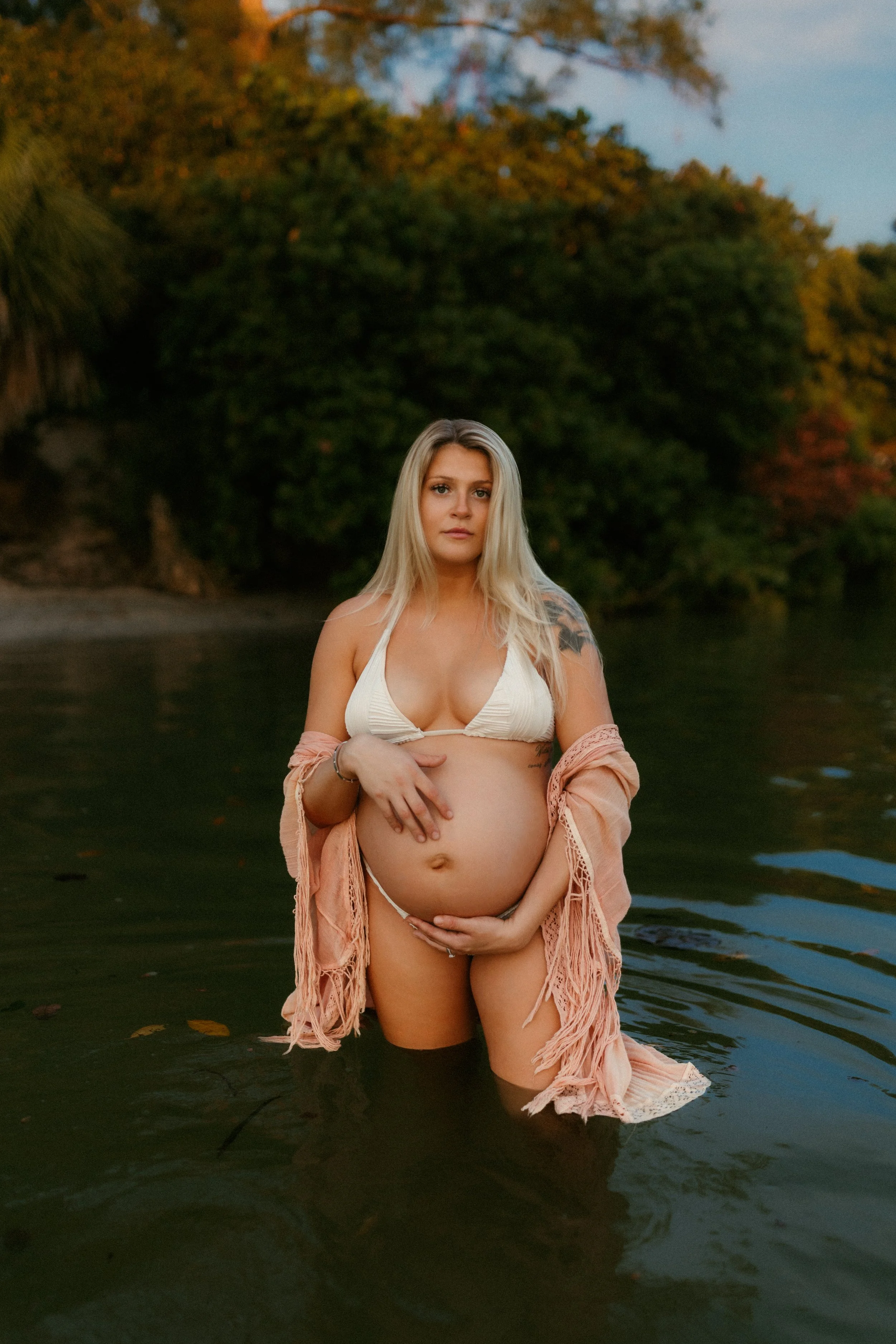 A pregnant woman standing in a body of water, wearing a bikini top and a fringed cover-up, with a scenic background of trees and a partly cloudy sky.