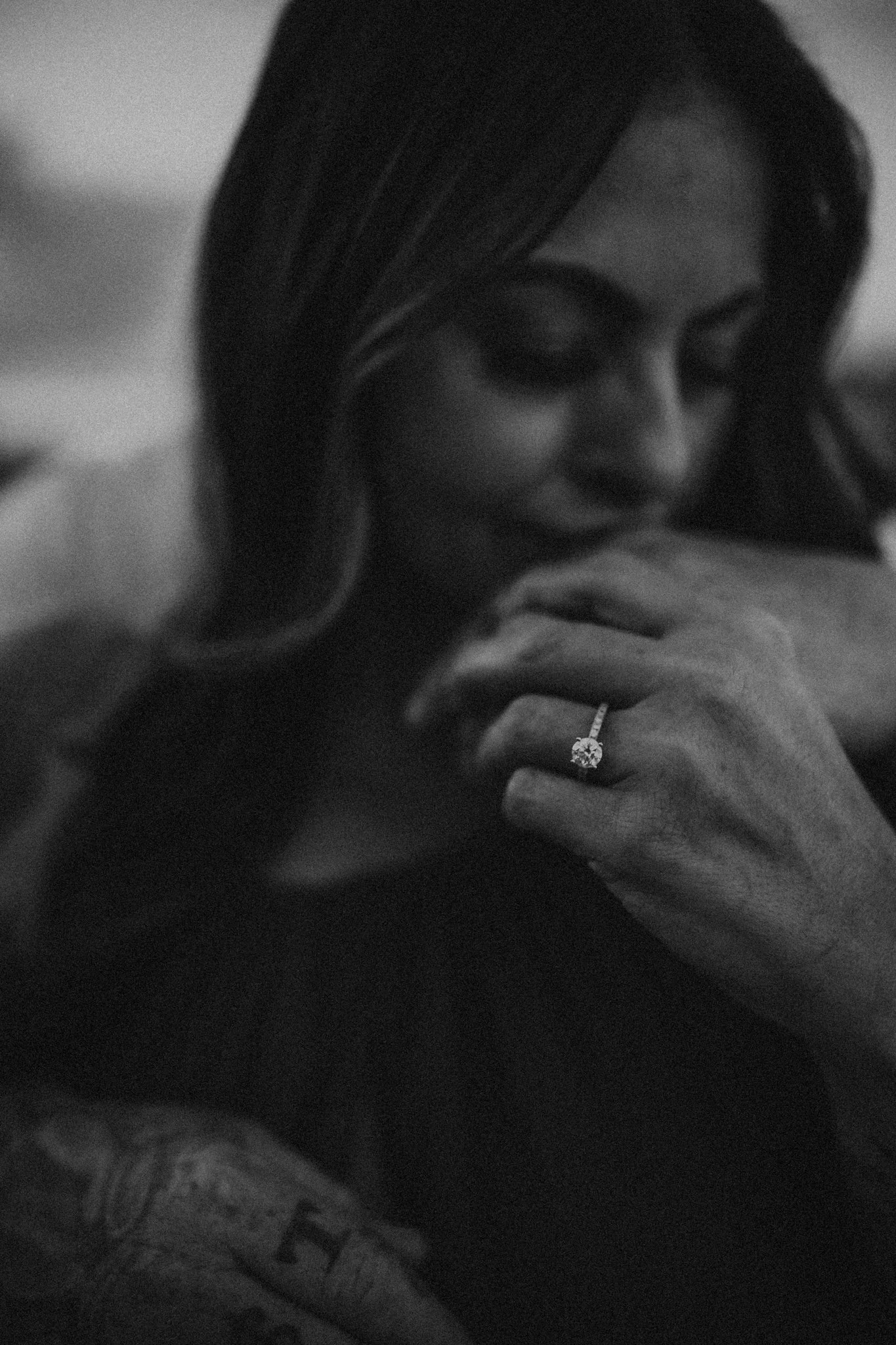 A woman with dark hair and makeup, looking down and touching her face. She is wearing a ring with a large gemstone and has tattoos on her arm, in black and white photography.