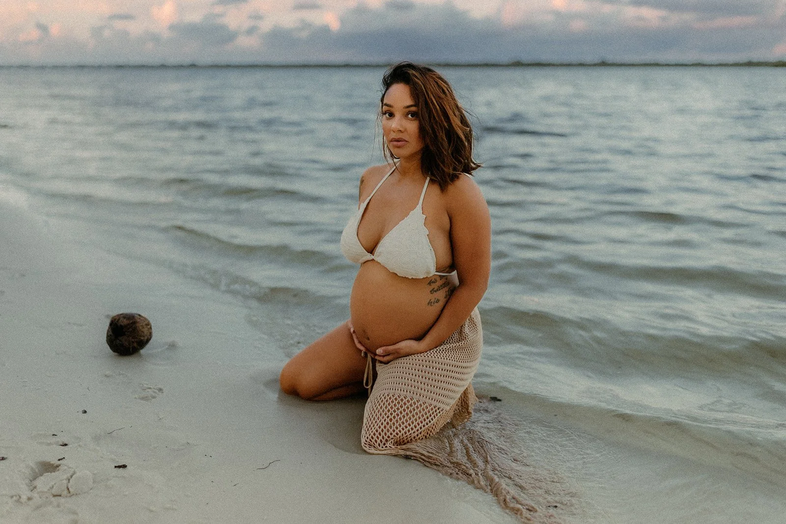 Pregnant woman kneeling on the beach near the water, wearing a beige bikini top and crochet skirt, with an expansive ocean view and cloudy sky in the background.