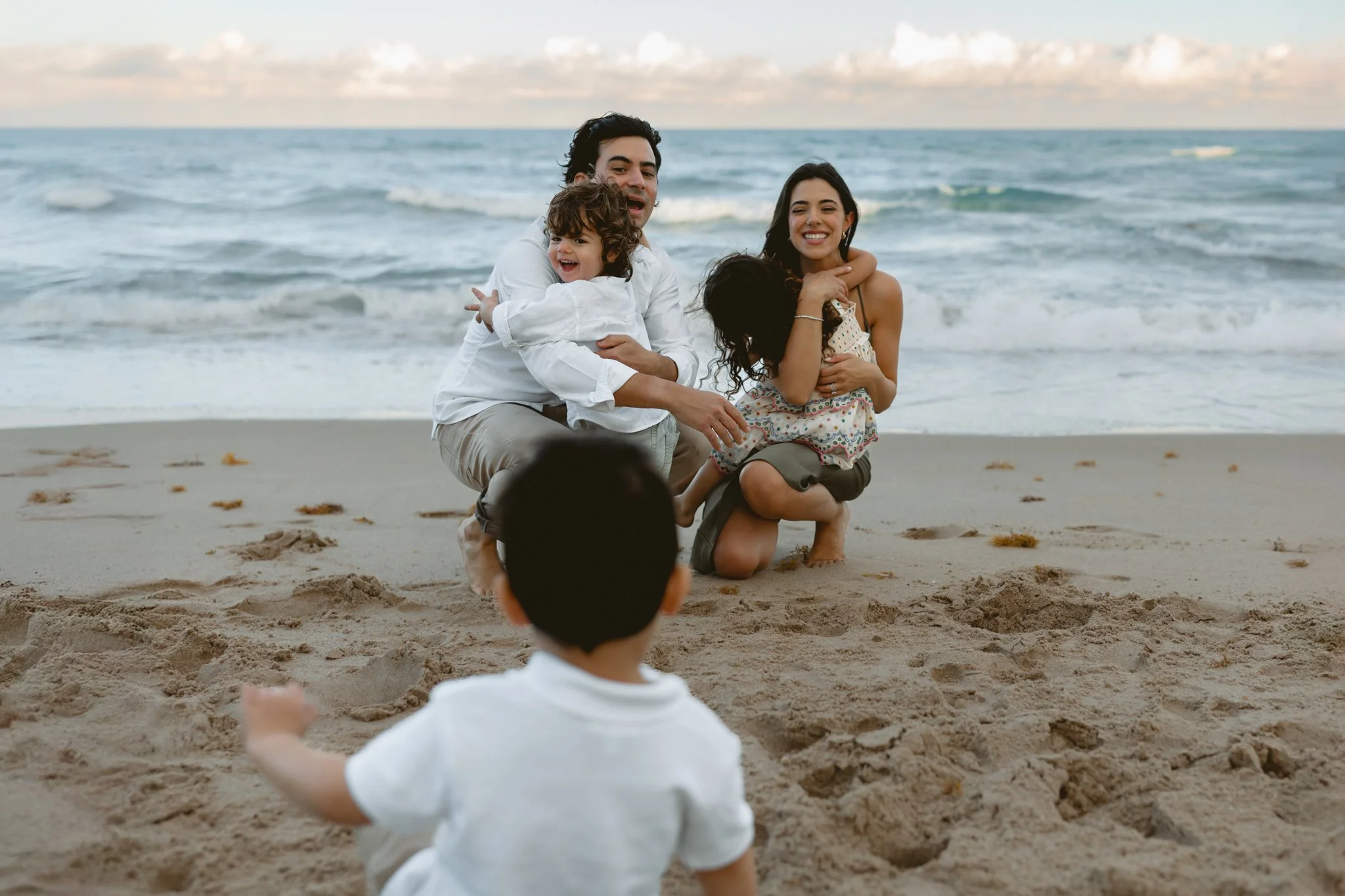 a mother and father kneeling down embracing their young children while looking at their young son on the beach