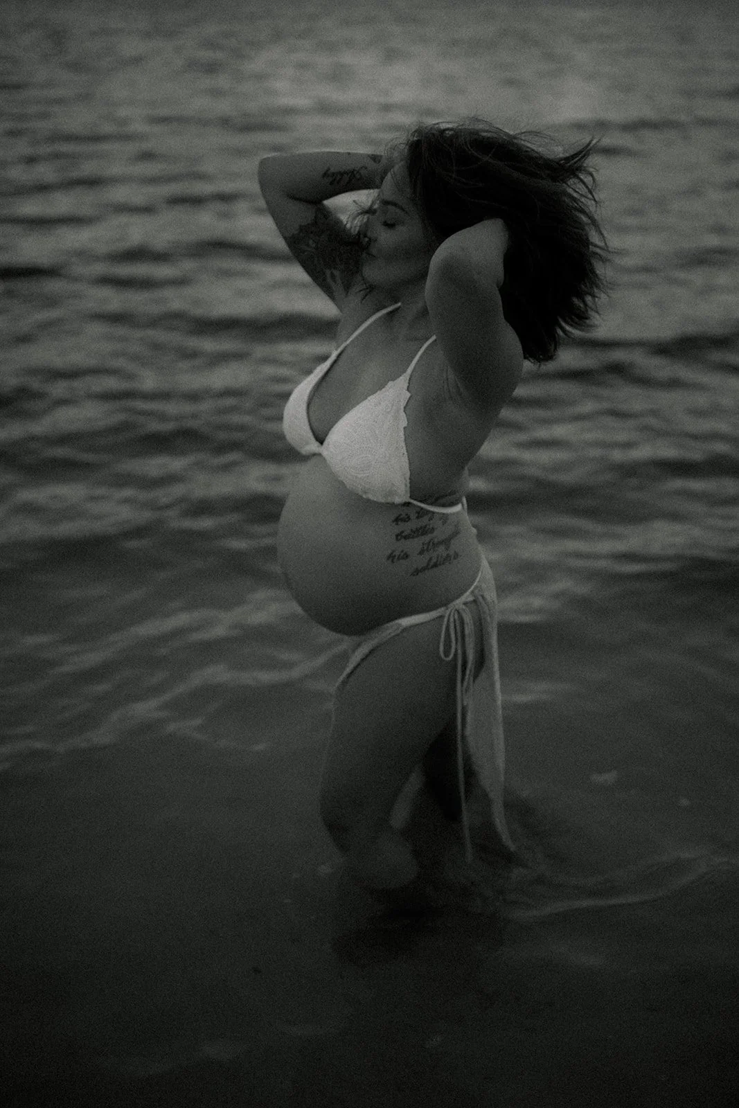 A pregnant woman standing in the ocean during sunset, wearing a bikini and with her hands behind her head.
