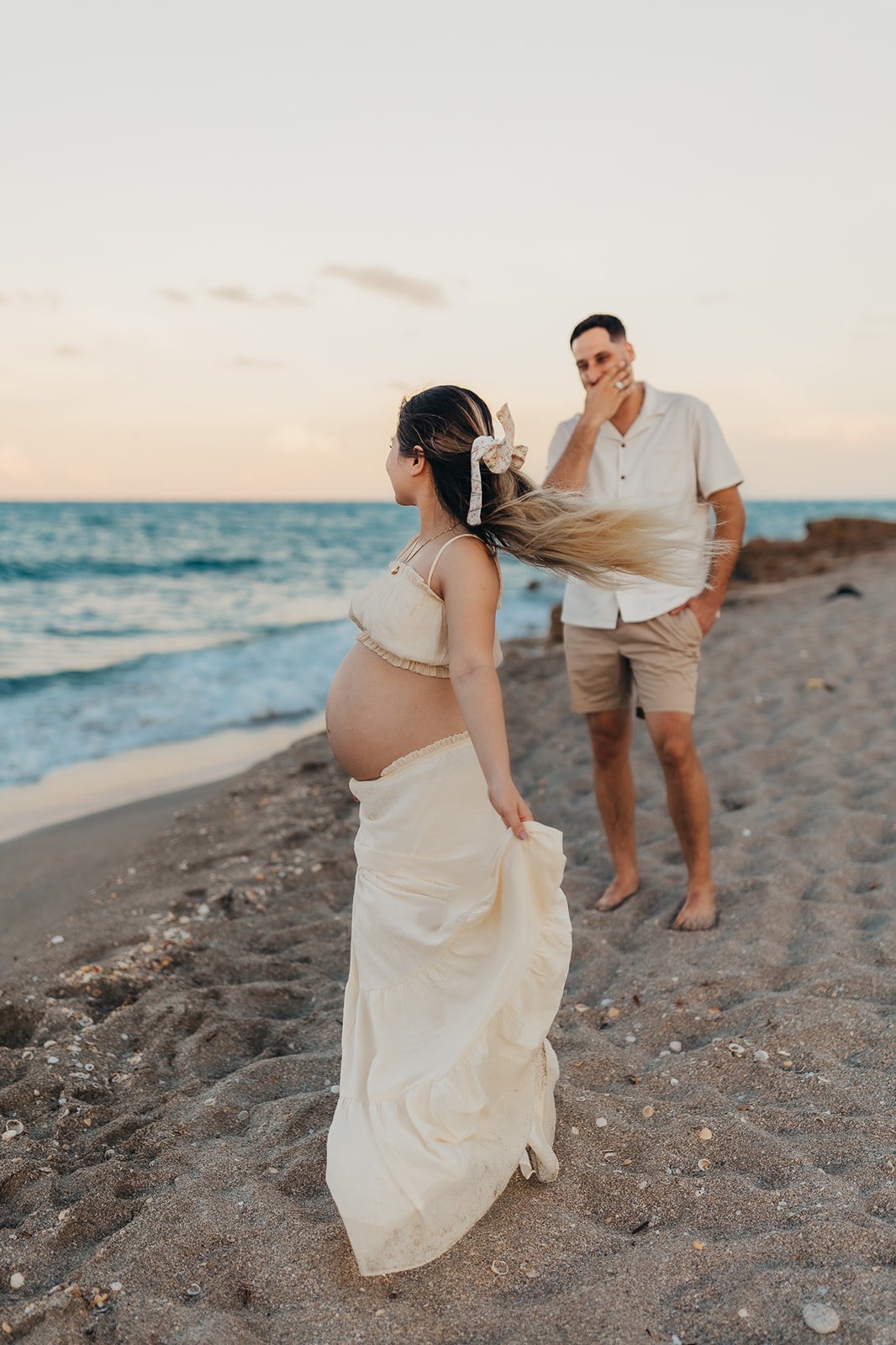A pregnant woman and a man standing on the beach, with the woman holding her dress and the man covering his mouth with his hand, during sunset.
