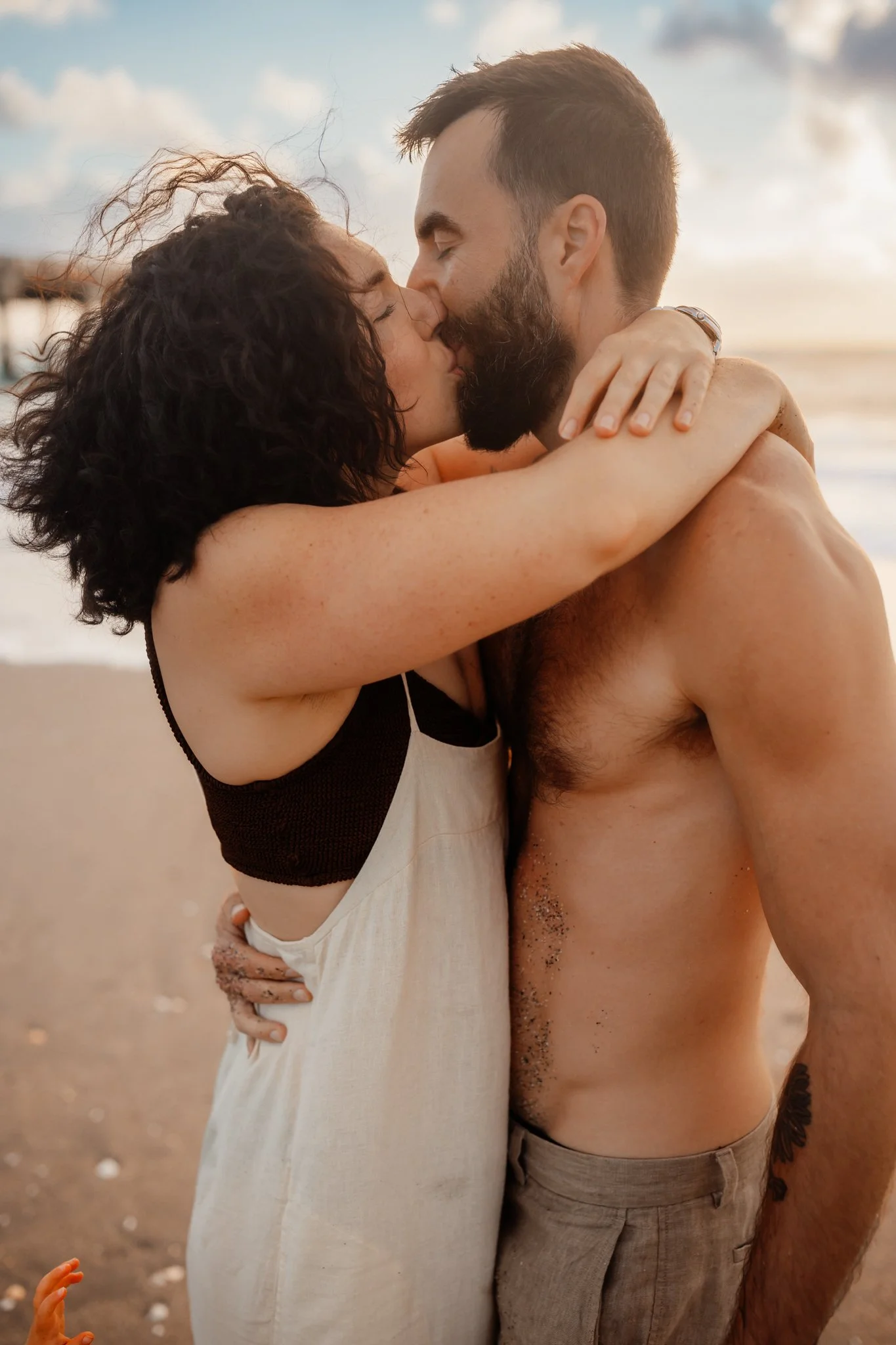 A couple kissing on the beach, embracing each other, with the ocean and sky in the background at sunset.