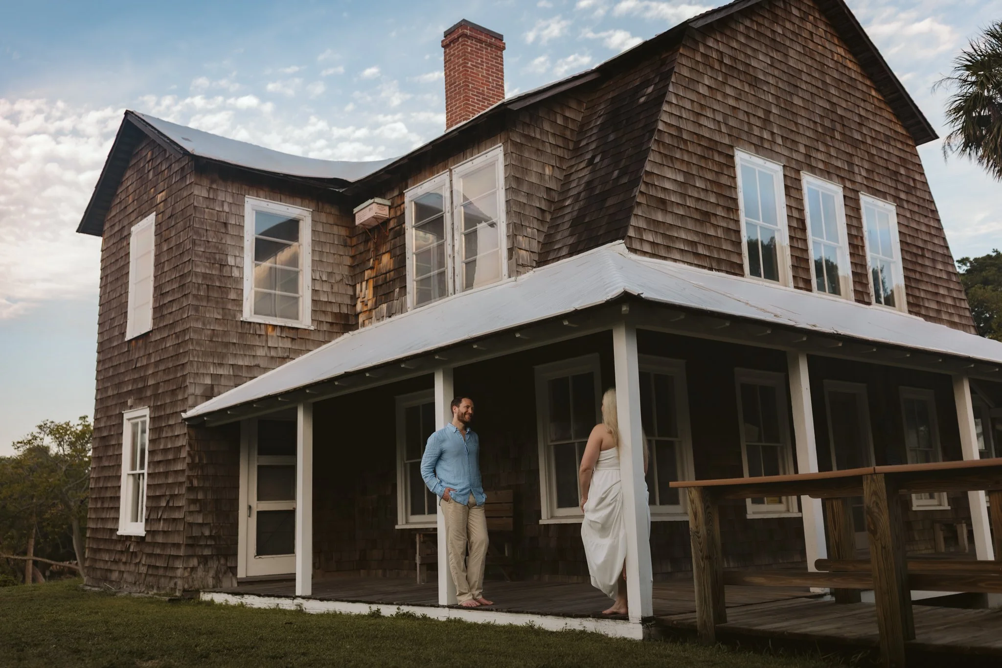A two-story wooden house with a porch, several windows, and a brick chimney. Two people, a man and a woman, are standing on the porch having a conversation. The sky is partly cloudy.
