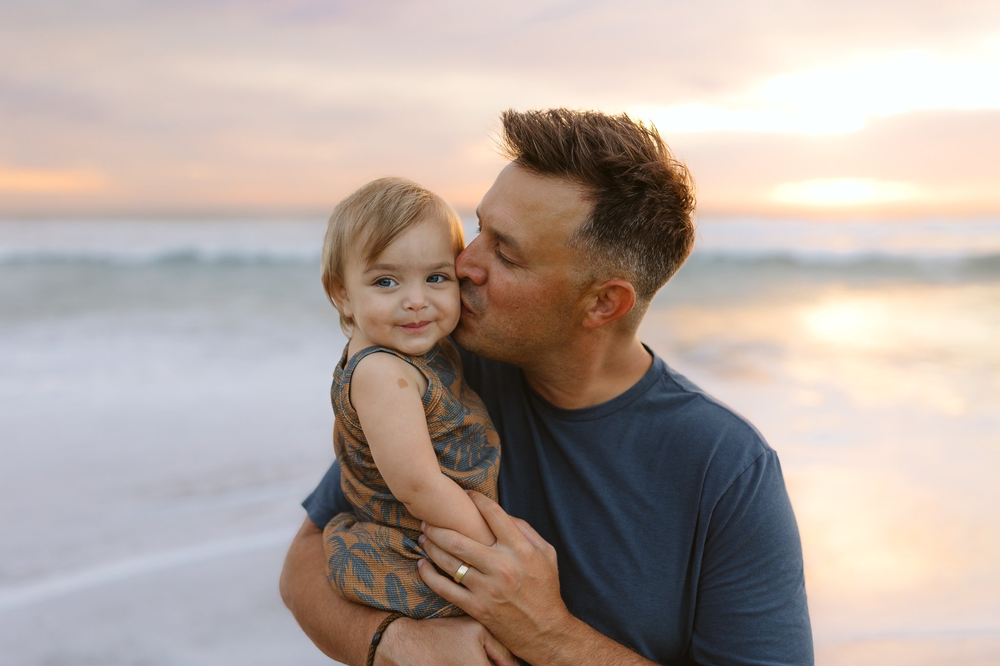 A man holding a young girl, kissing her cheek, on the beach at sunset.