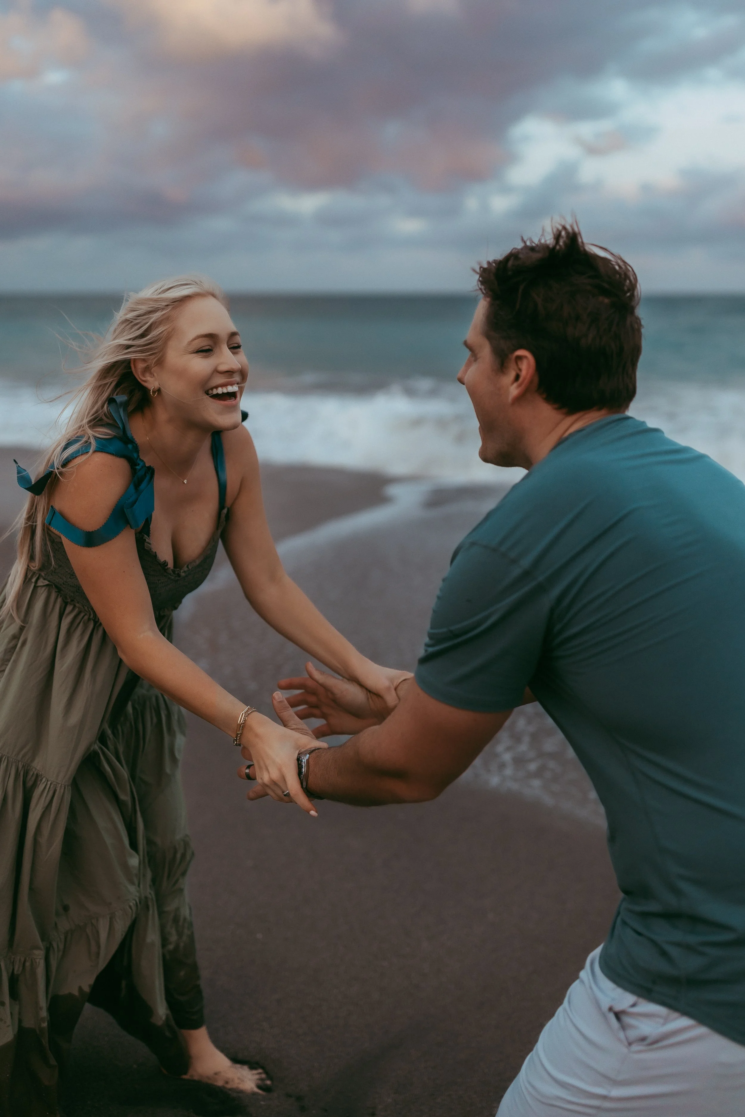 A happy woman is at the beach, laughing and holding hands with a man, with the ocean and cloudy sky in the background.