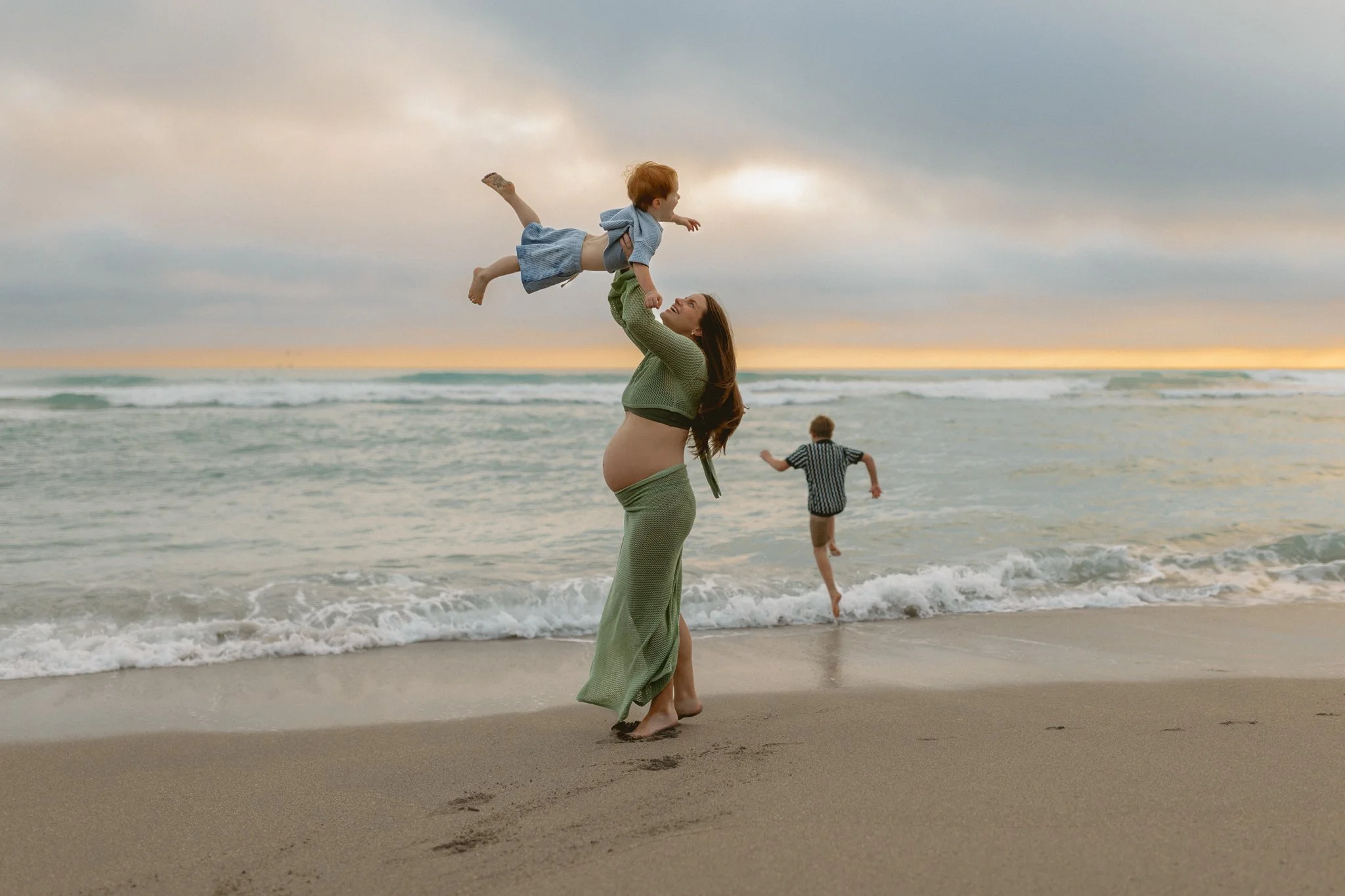 a pregnant woman lifting up her son in the air on the beach while her other son is running towards the ocean