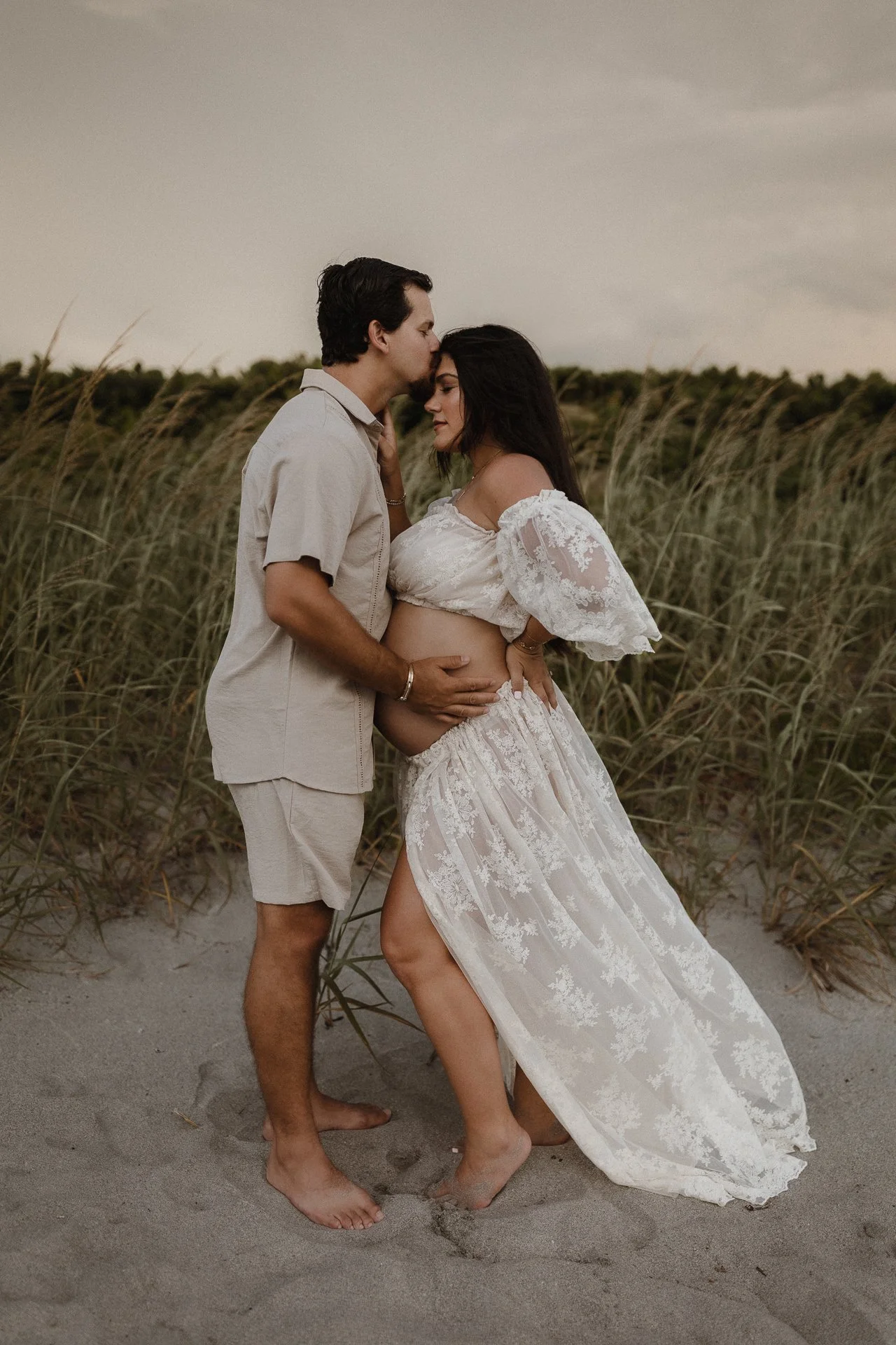 A couple standing on a sandy beach with tall grass in the background, with the man gently holding the woman's baby bump as she leans into him, dressed in a white floral skirt and off-shoulder top, during sunset.