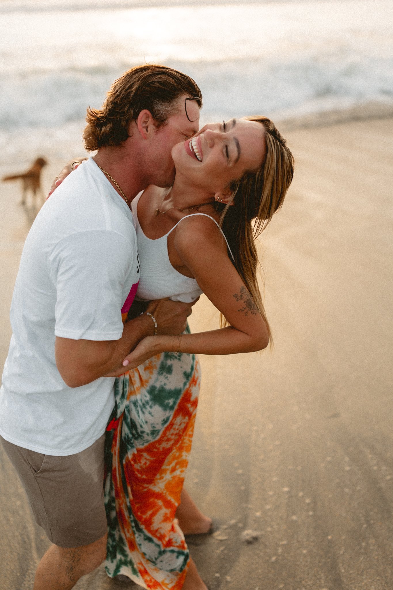 A young couple is laughing and playing on the beach, with a dog in the background during sunset.