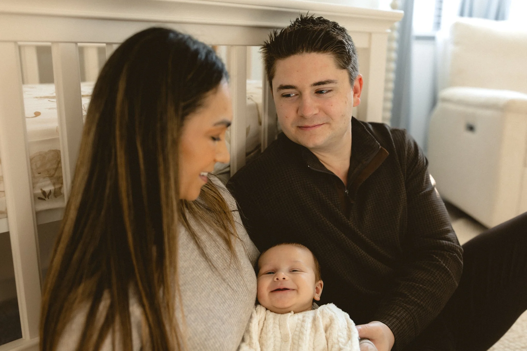 A family sitting together with a mother holding a smiling baby, a father looking at the baby and mother, in a cozy room with a crib and a small air purifier in the background.