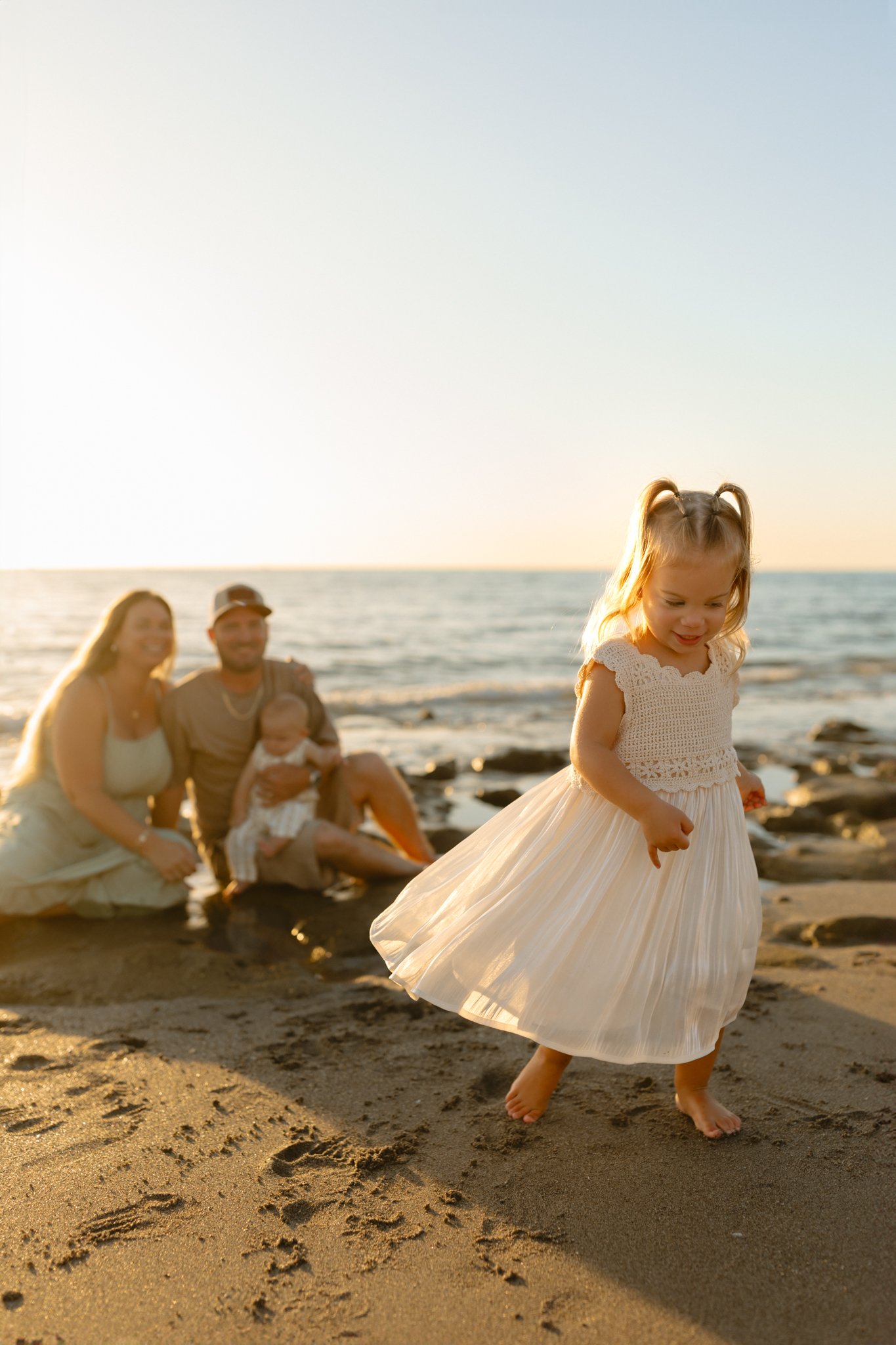 A young girl in a white dress is walking barefoot on the beach with a family sitting in the background near the water at sunset.