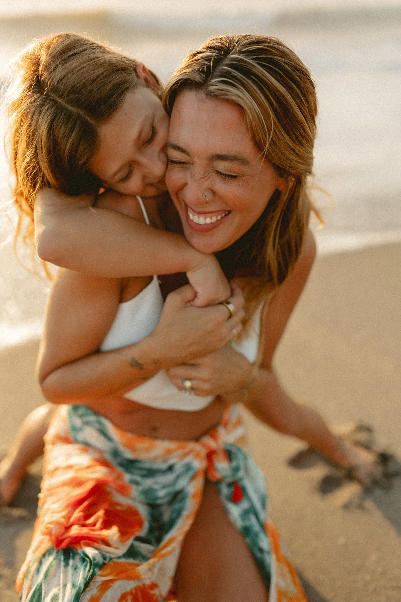 A woman with a nose piercing and a smile, embracing a young girl on a sandy beach during sunset.