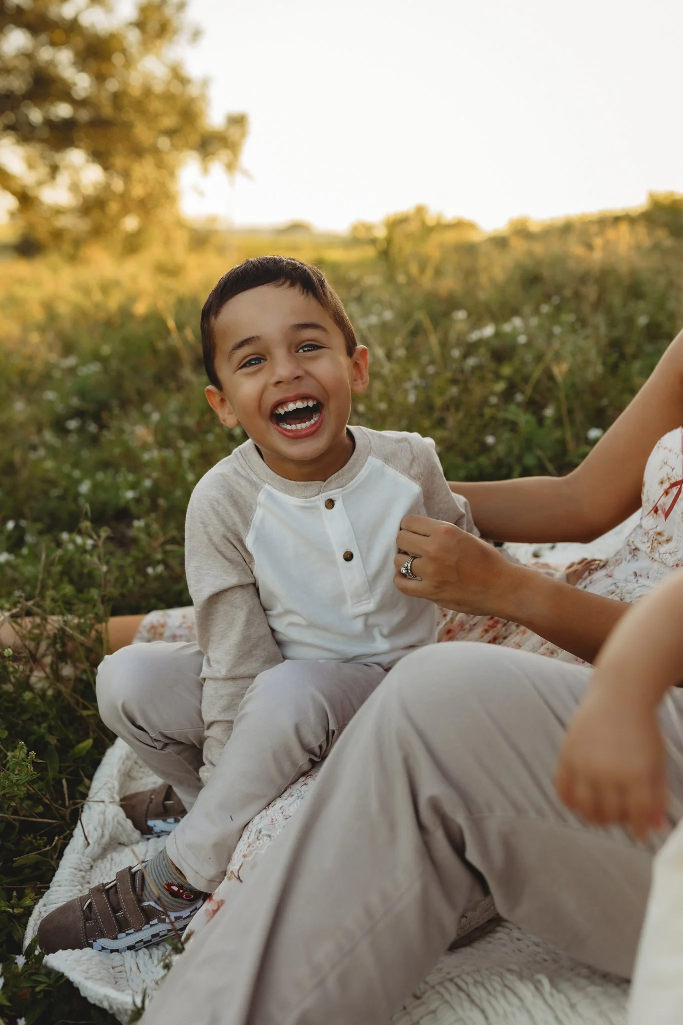 A young boy sitting on a blanket outdoors, smiling and laughing, with a woman next to him in a field with green plants and trees in the background.
