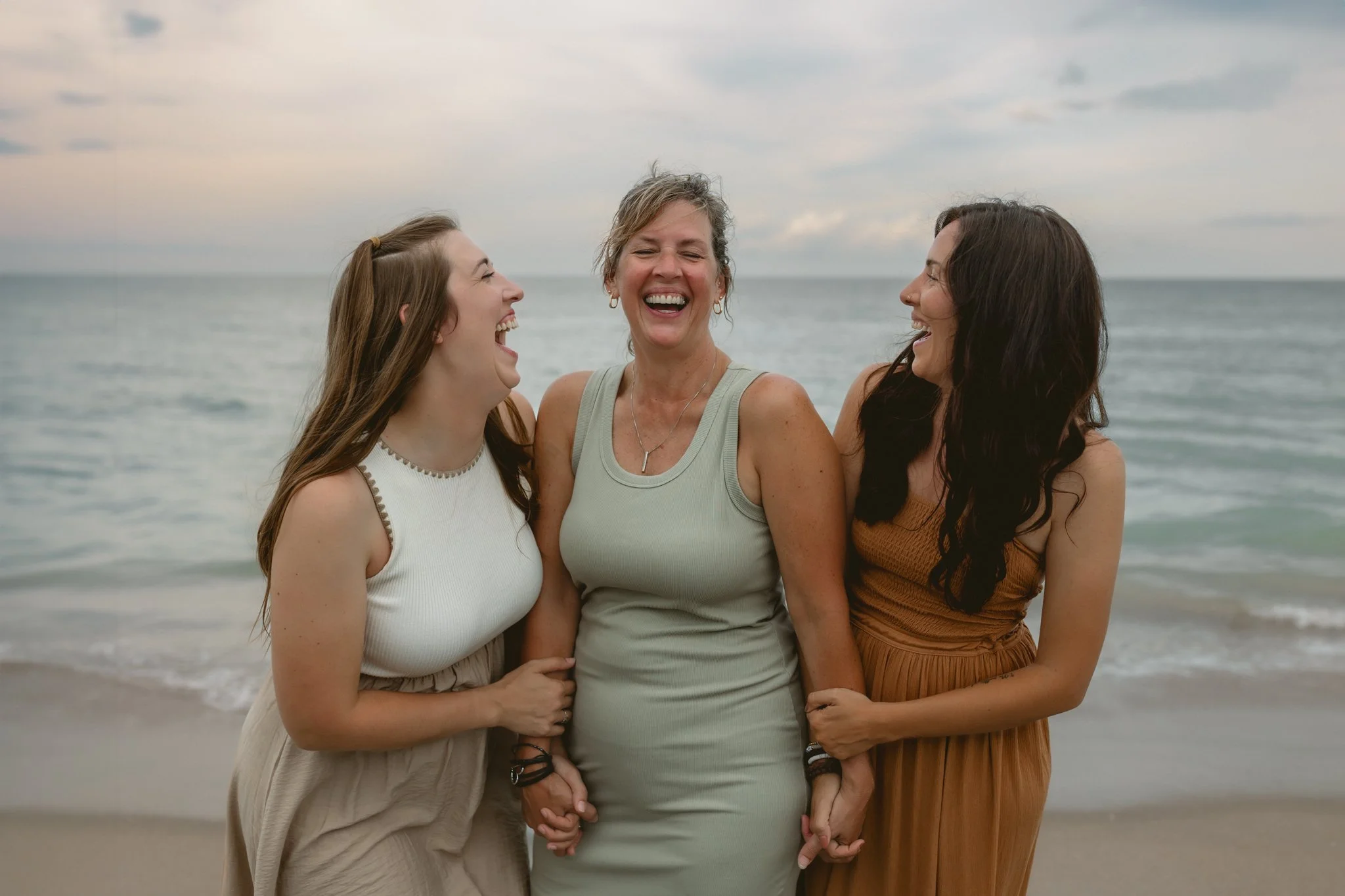 Three women in beachwear laughing and holding hands on the beach during sunset.