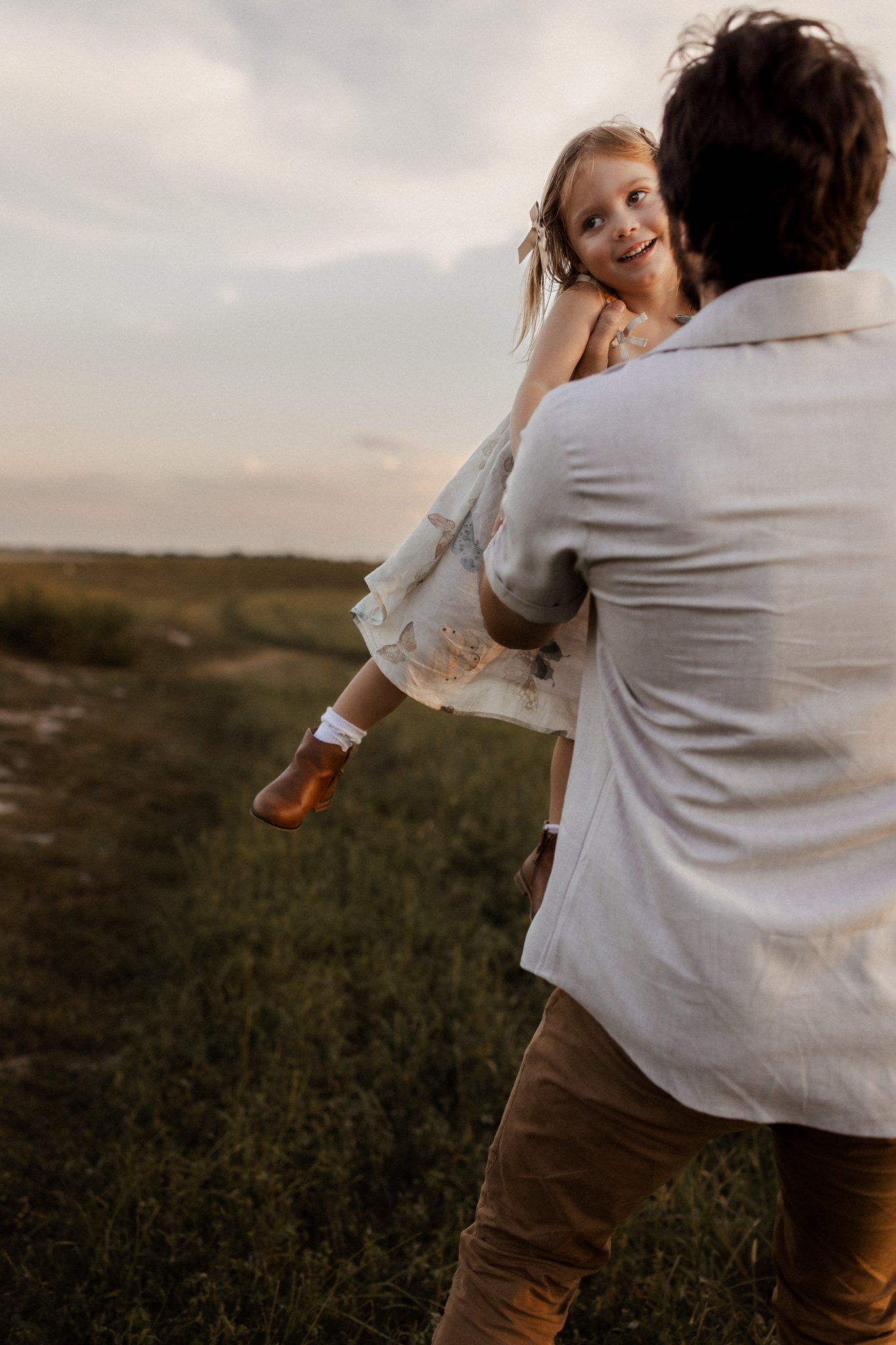 A man is holding a young girl in the air outdoors during sunset, with a grassy landscape in the background.