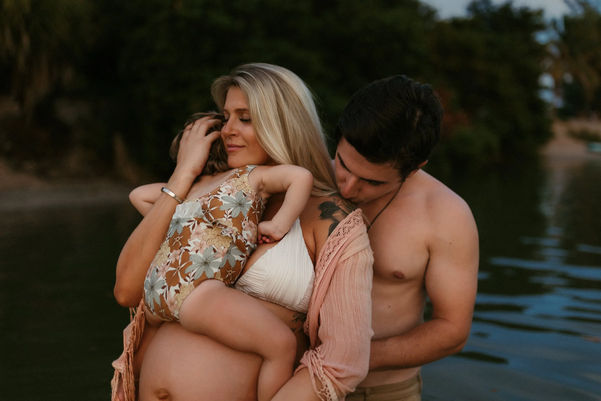A woman holding a young girl in her arms while a shirtless man kisses her shoulder, near a body of water at sunset.