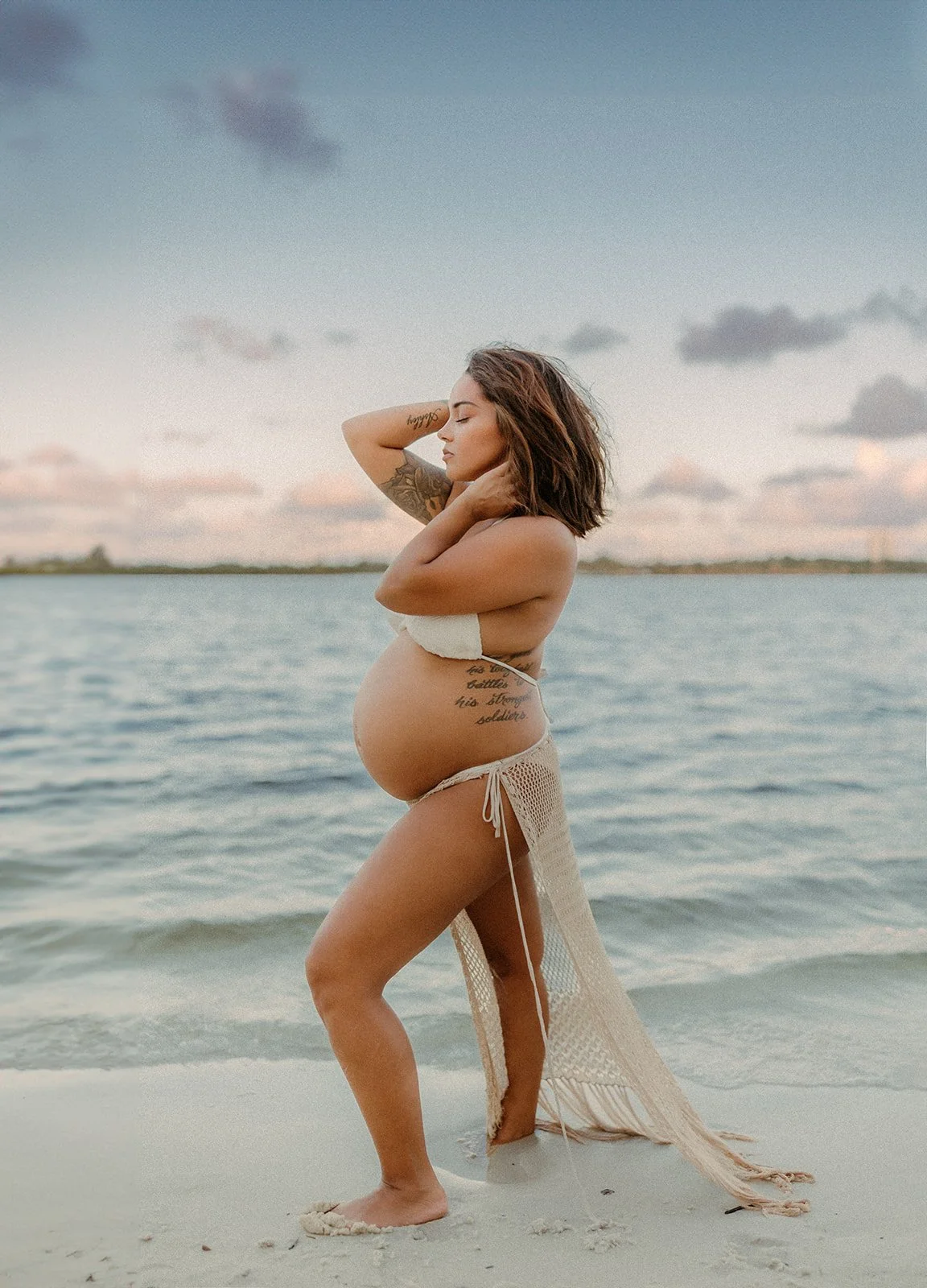 Pregnant woman standing on the beach near water, wearing a bikini and a crocheted cover-up, with her hands behind her head and eyes closed, during sunset.
