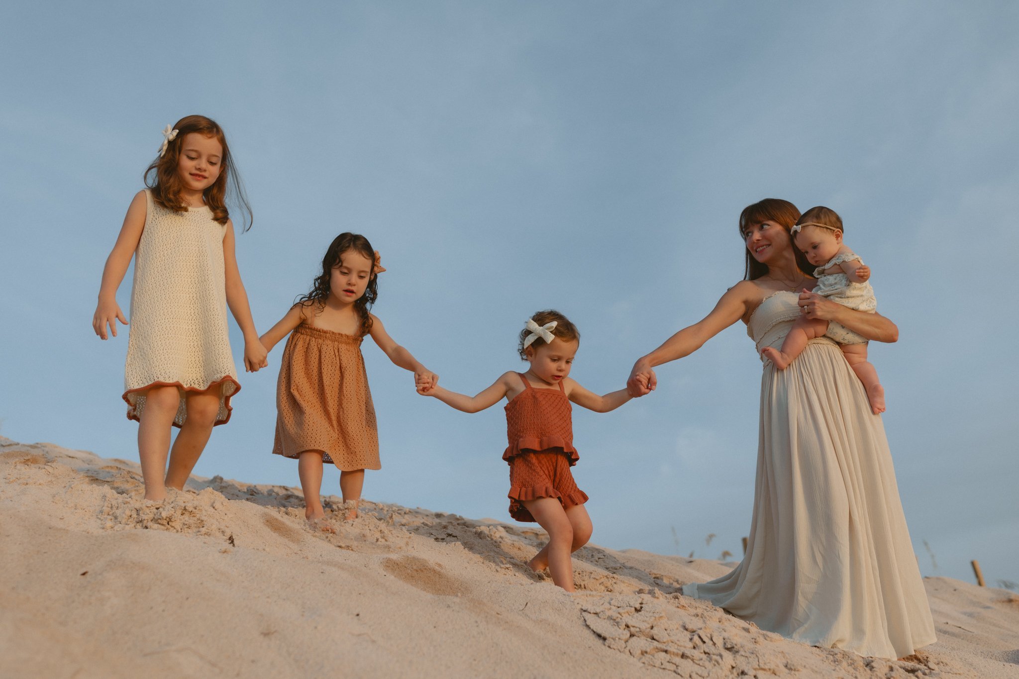 A woman and four young girls holding hands walk down a sandy hill at the beach during sunset.