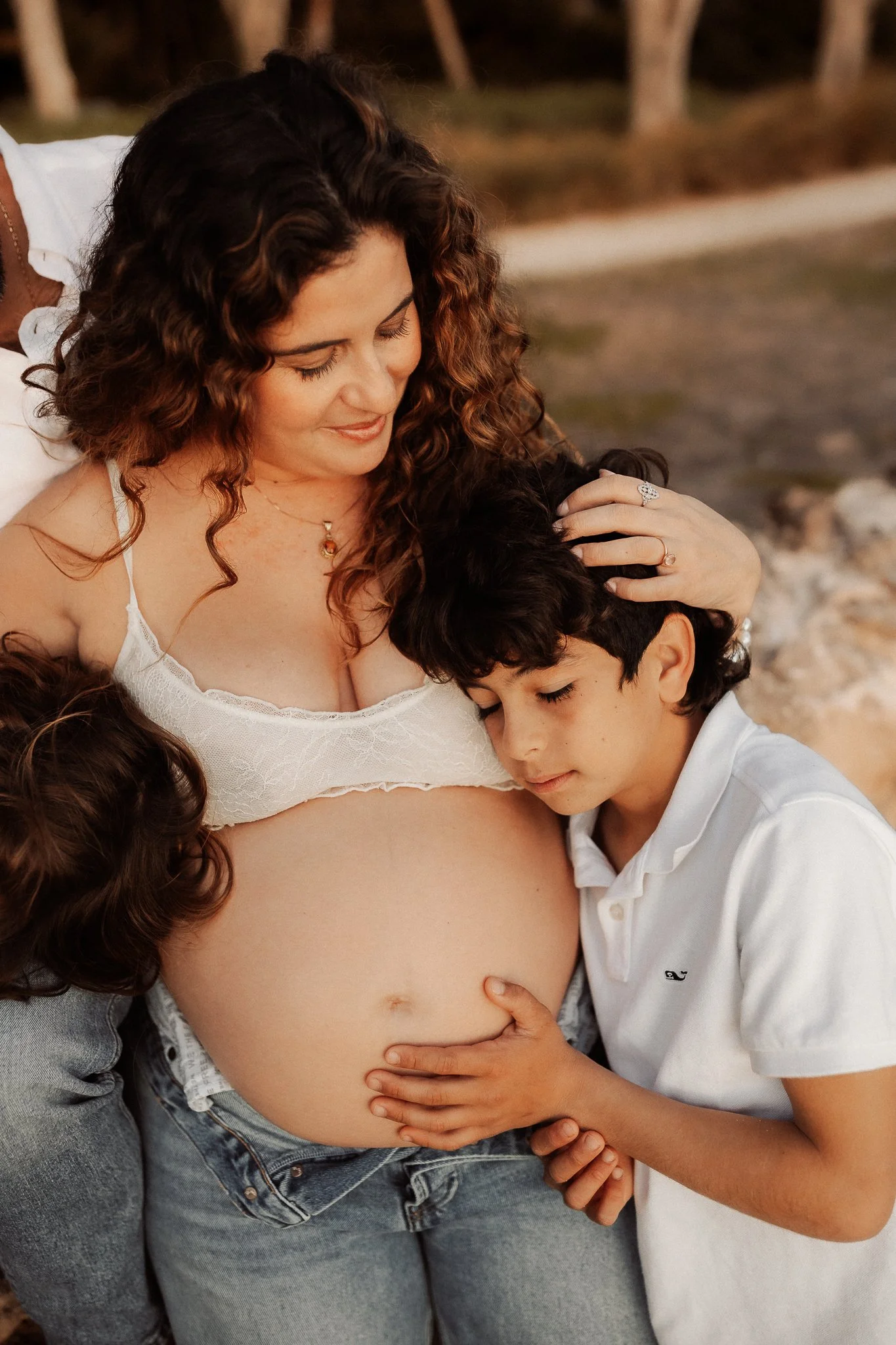A pregnant woman with curly brown hair in a white top, surrounded by children, touching her belly, outdoors with trees in the background.