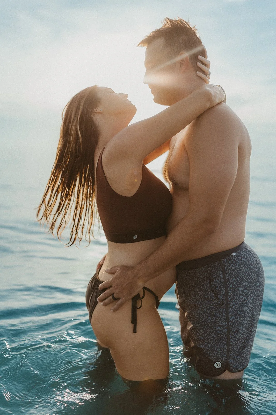 A man and woman embracing in water at a beach during sunset, with the sun's rays creating a glow around them.