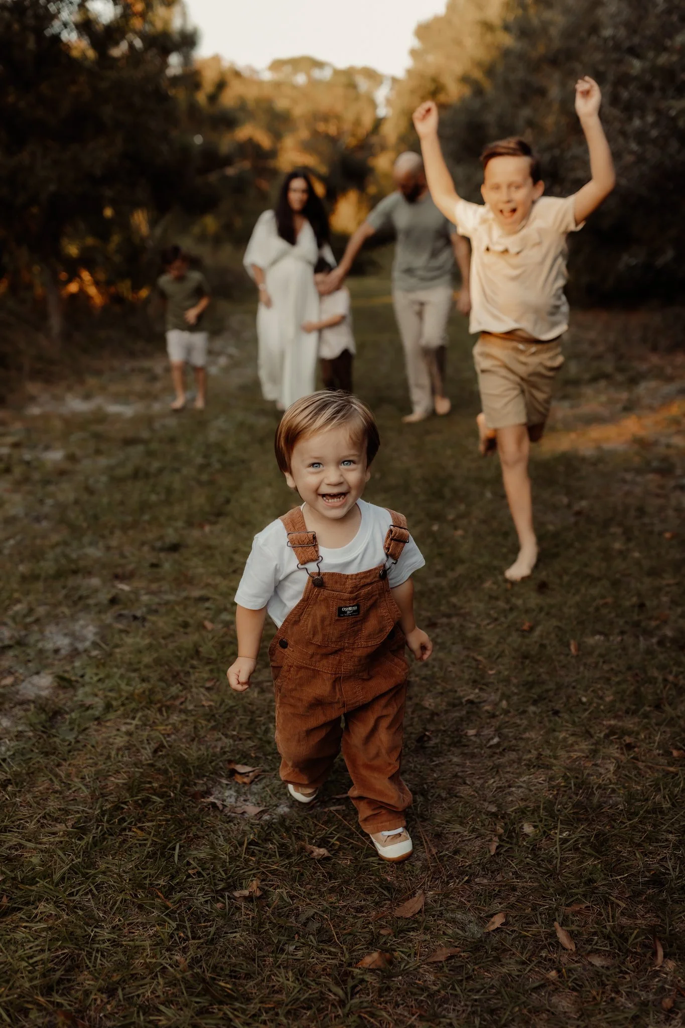 A young boy with brown hair, wearing a white shirt and brown overalls, happily running on a grassy path outdoors while a group of family members, including a man, woman, and children, cheer and chase him in the background during the daytime.