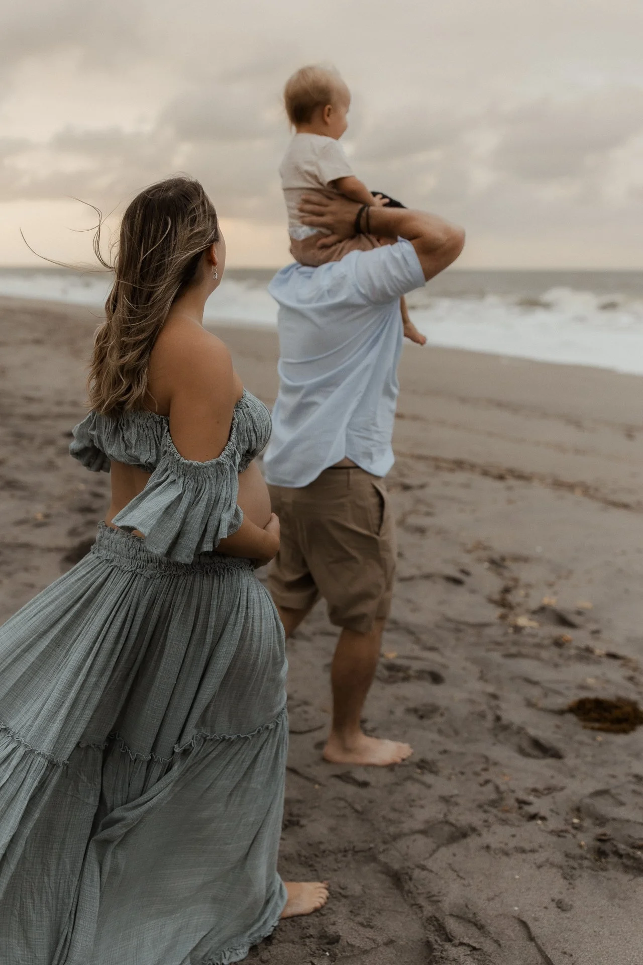 A man carrying a child on his shoulders on a beach during sunset, with a woman standing nearby, all looking towards the ocean.
