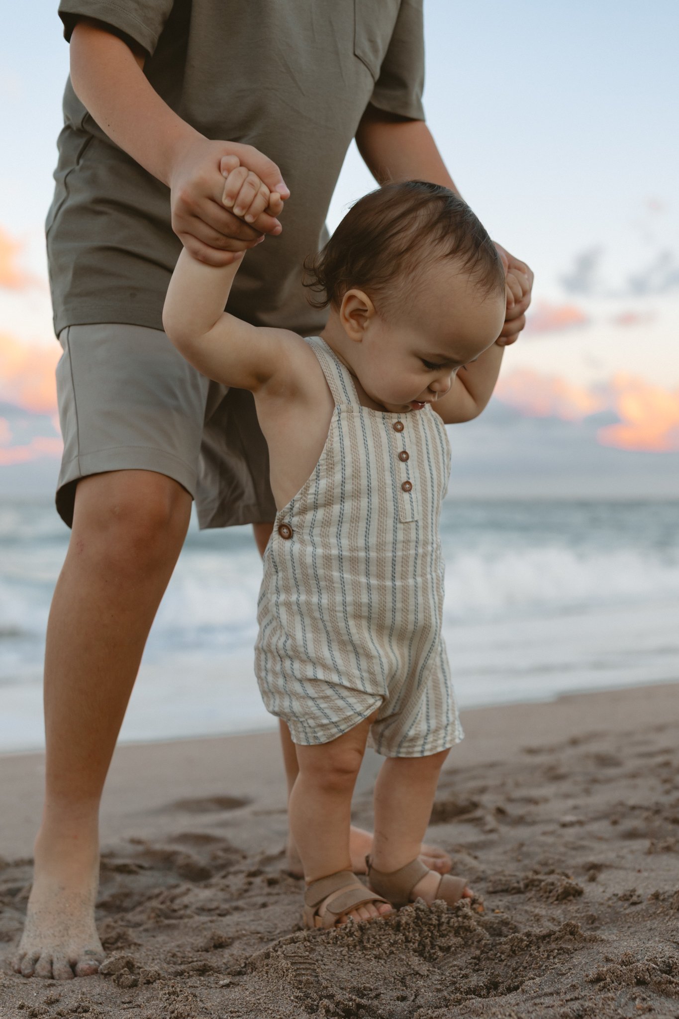 A young child learning to walk on the beach with help from an adult, holding the child's hands. The child is wearing a striped romper and sandals, and the adult is barefoot, standing in sand near the ocean, with a sunset sky in the background.