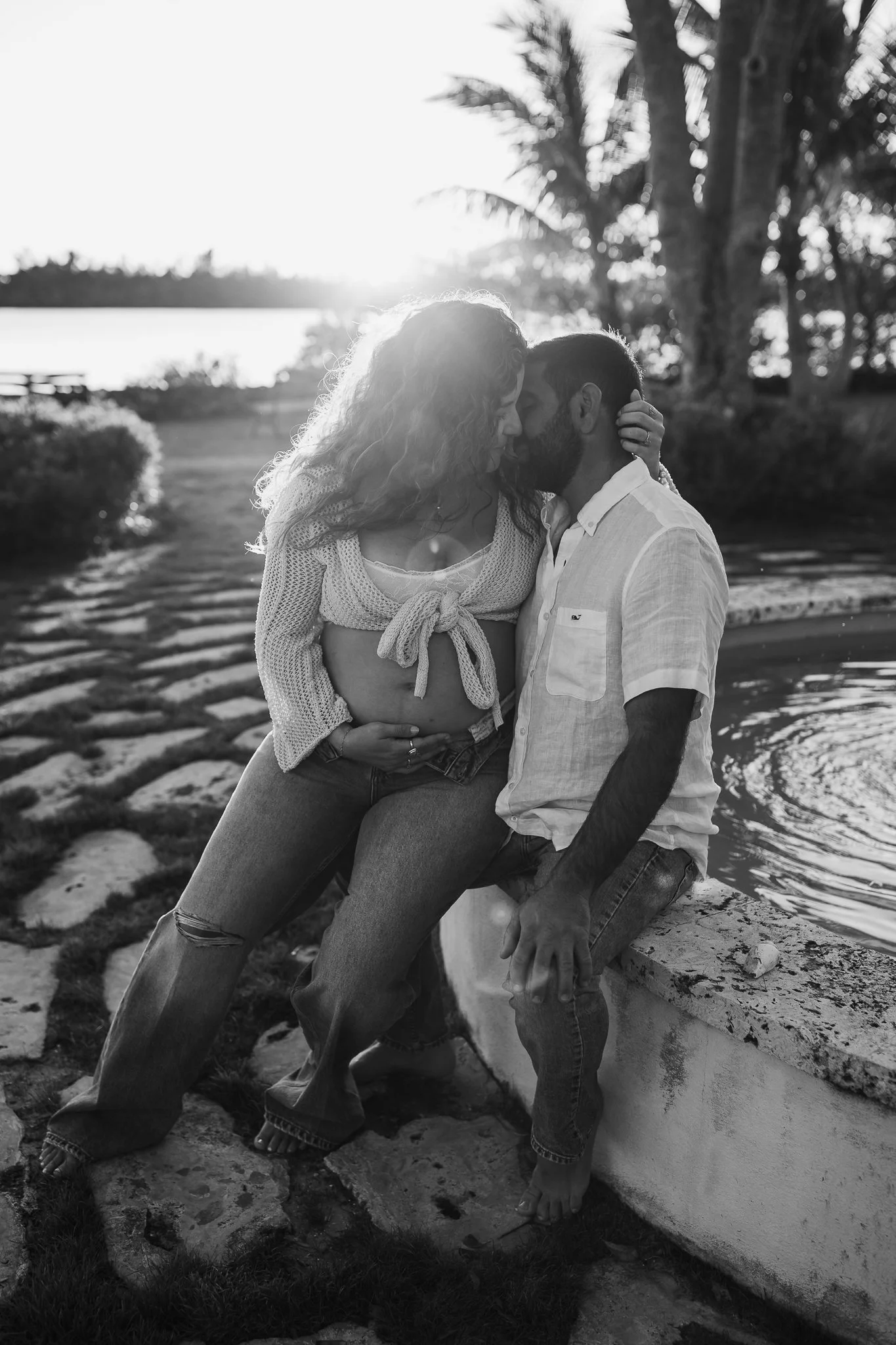 A black and white photo of a pregnant woman and a man sharing a close moment outdoors near water during sunset, with palm trees in the background.