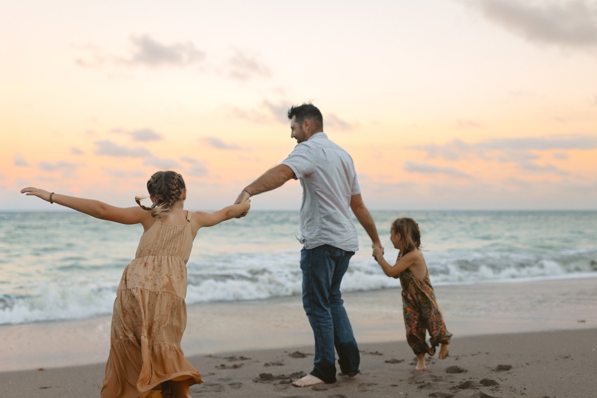 A man playing with two young girls on the beach at sunset, holding their hands as they walk near the water.