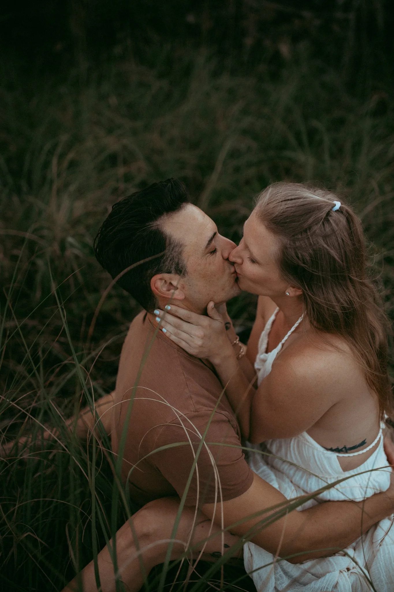 A couple sitting among tall grass, kissing passionately.