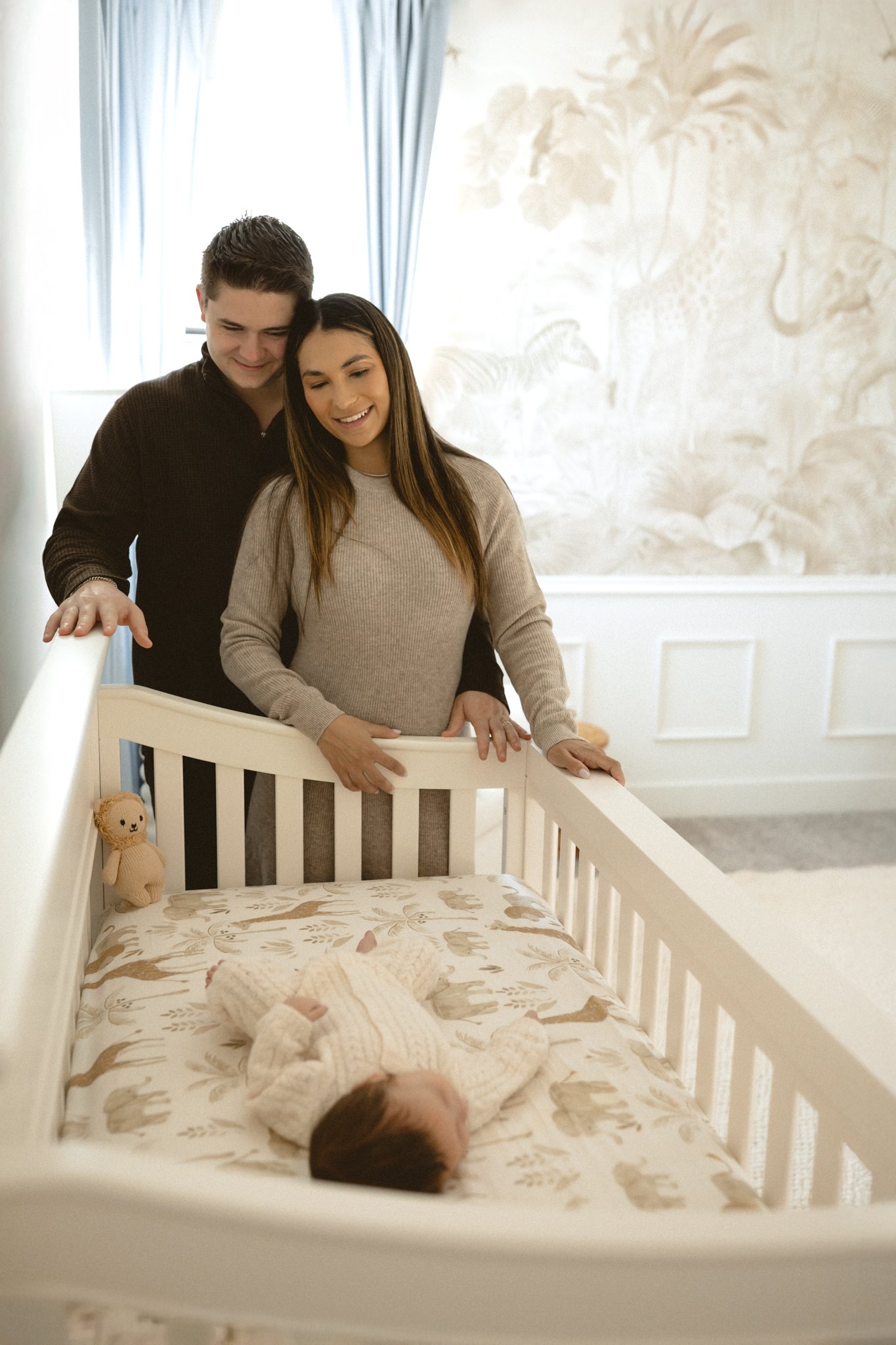 A couple, a man and a woman, standing next to a crib, looking at a sleeping baby inside, with a stuffed toy resting on the crib rail.