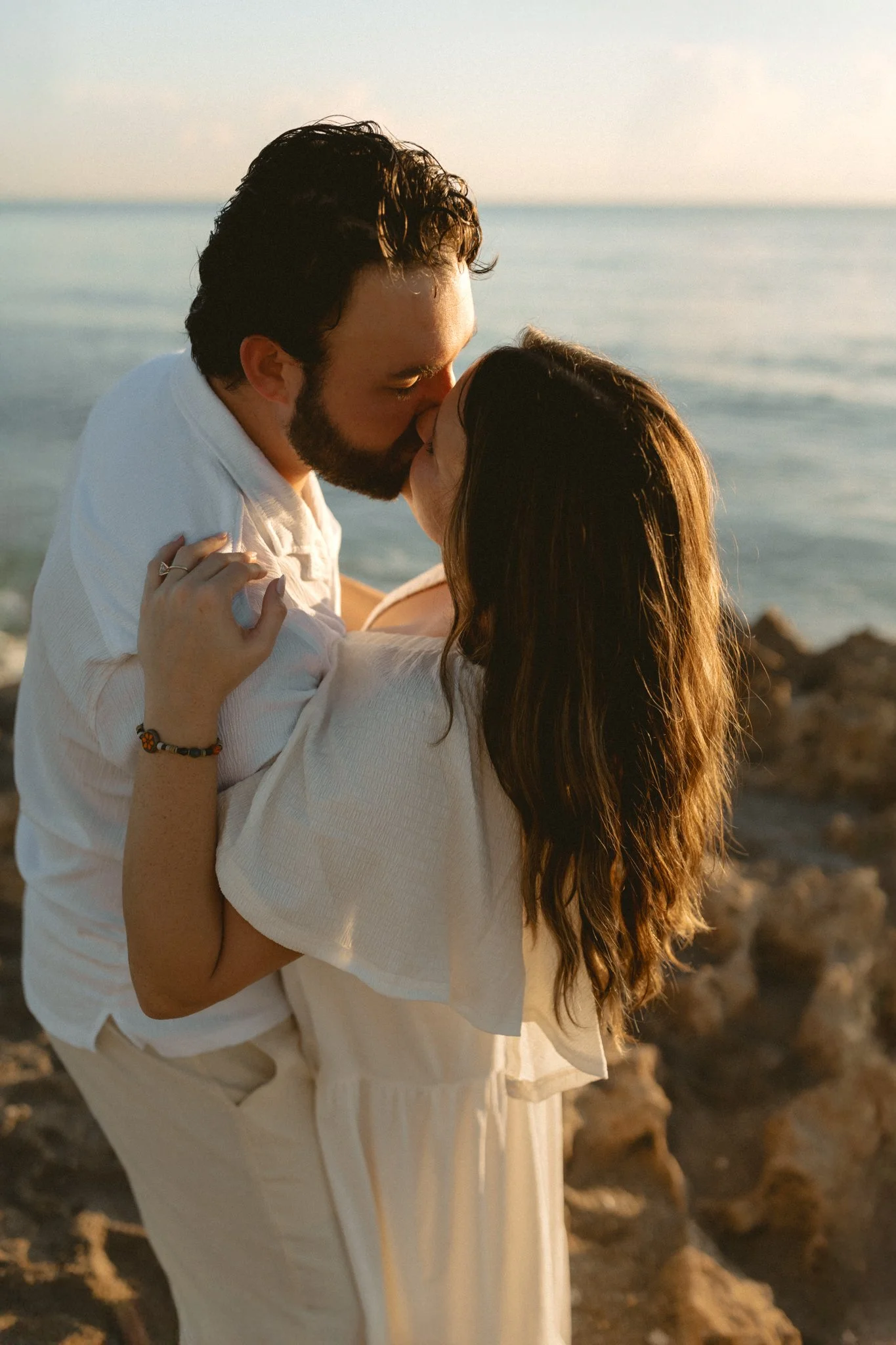 A couple sharing a kiss on a beach during sunset, with rocks and the ocean in the background.