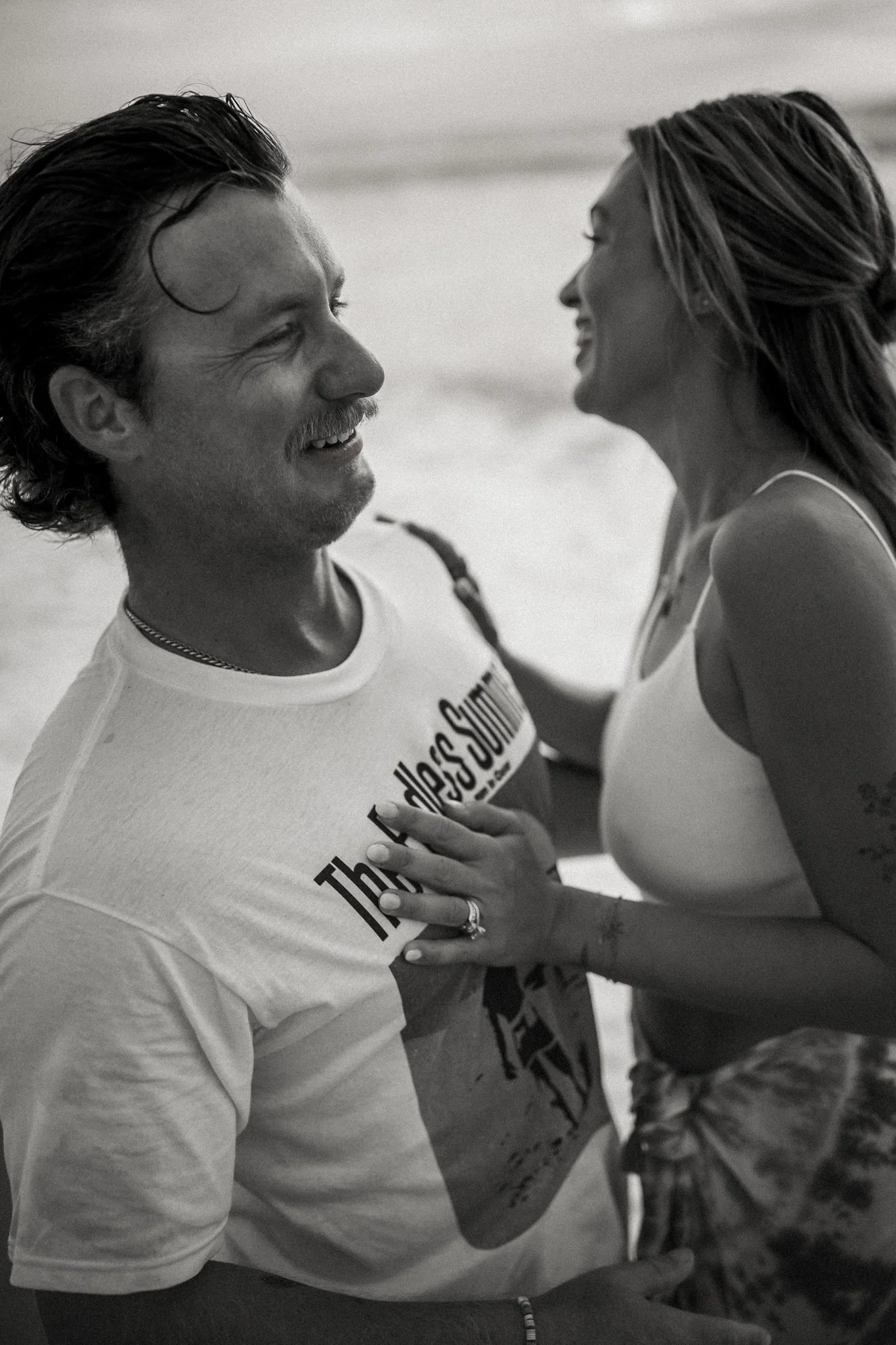 A black and white photo of a man and a woman smiling and looking at each other on the beach, with the woman touching the man's chest.