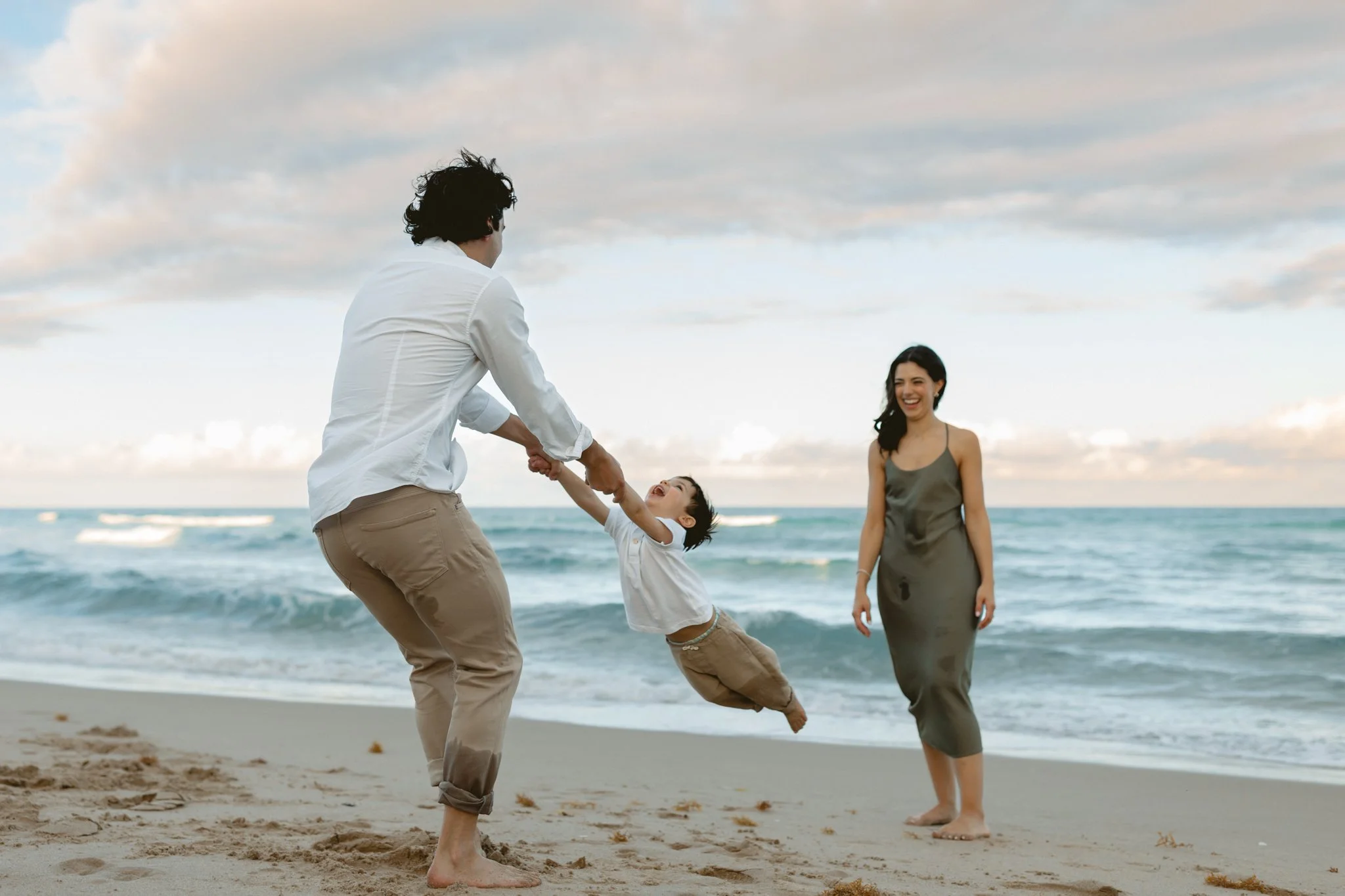 father swinging young son on the beach with mom laughing in the background