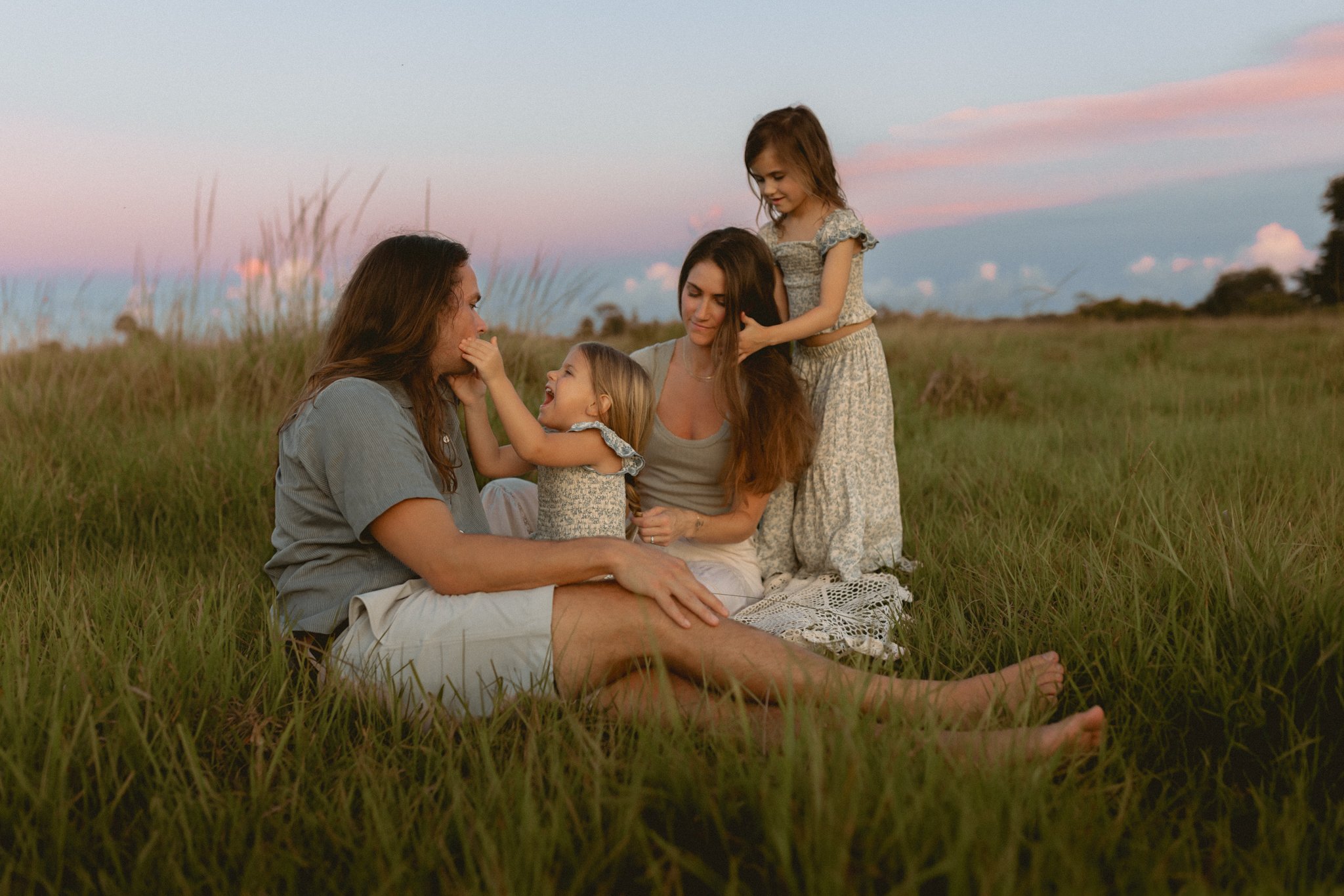 Family of four, two women and two young girls, sitting in a grassy field during sunset, playing and smiling together.