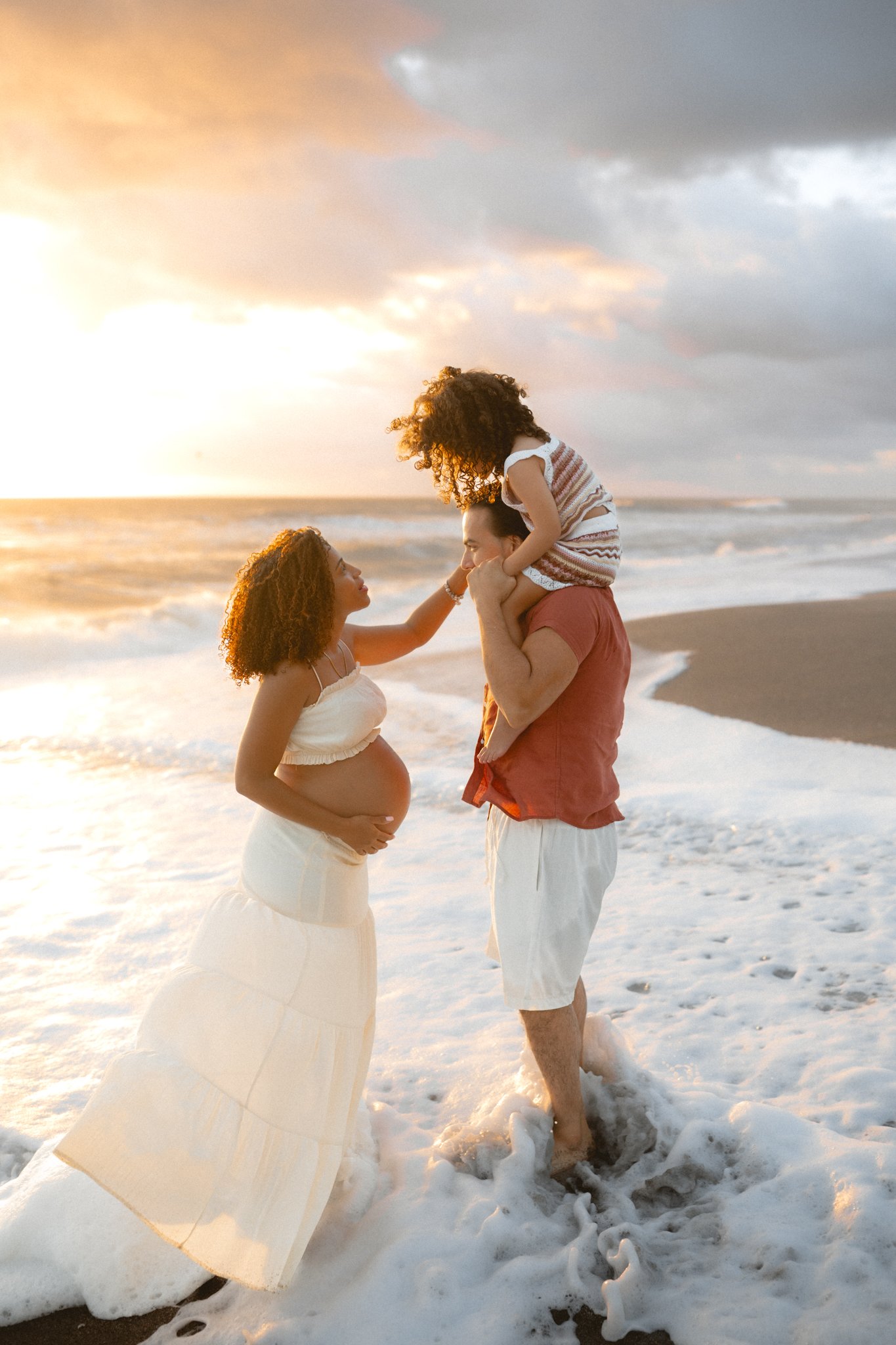 Family at the beach during sunset, with a pregnant woman, a man holding a child on his shoulders, and a woman touching the child's face.