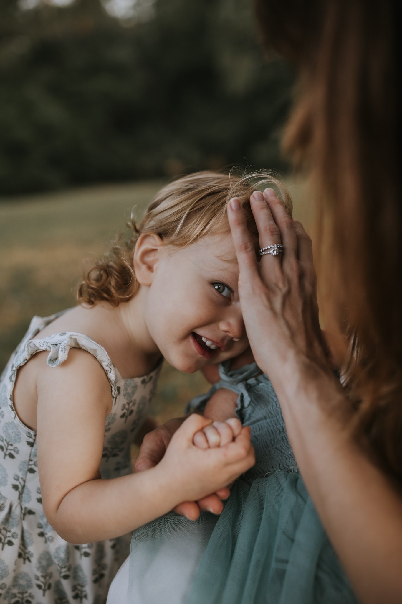 A young girl plays a game with an adult, both smiling, outdoors in a park or garden, the girl has blonde hair and wears a patterned dress, the adult has reddish hair and is wearing a light blue top.