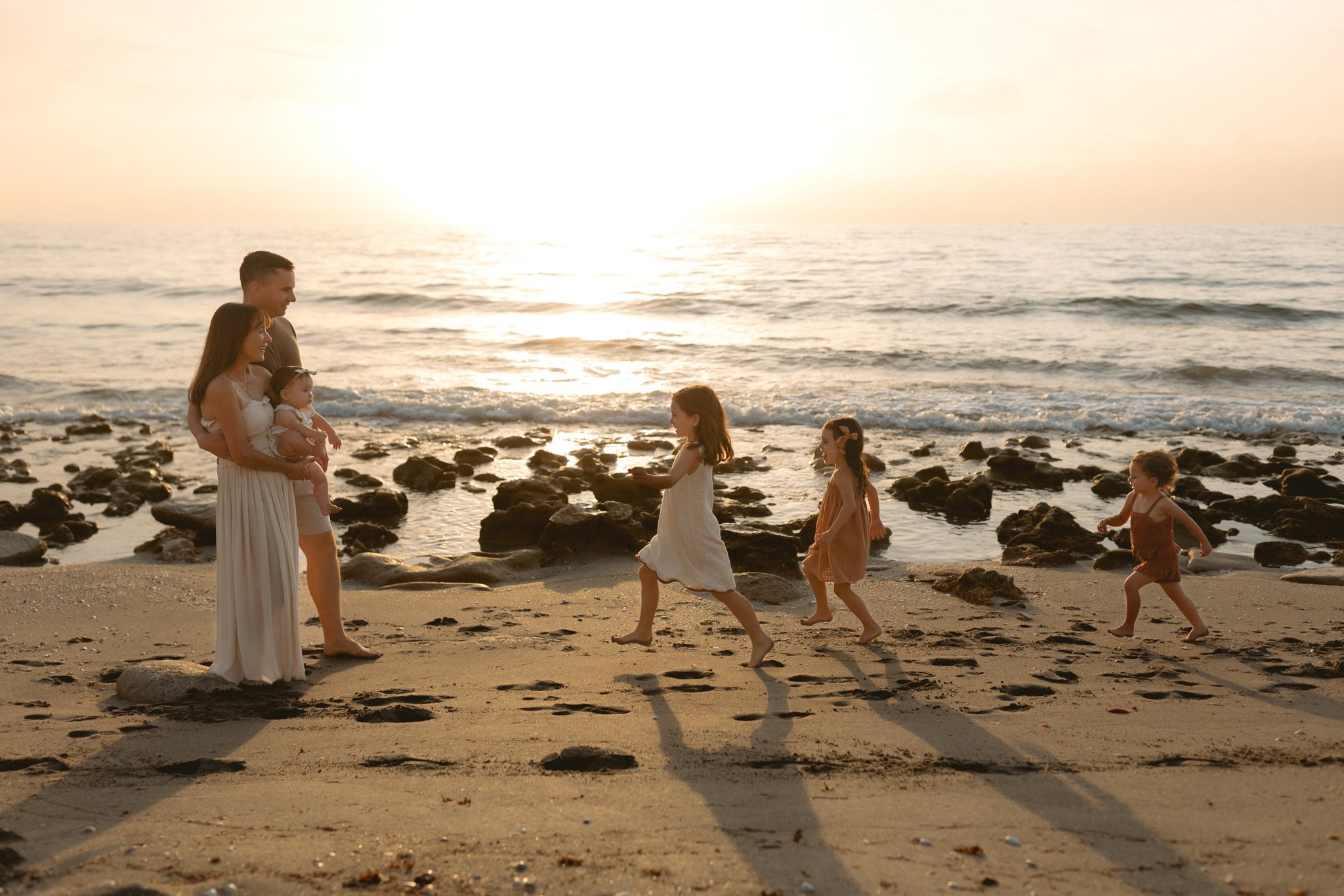 A family of six, including a mother, father, and four children, enjoying a sunset on a rocky beach. The mother is holding a baby, while the children are running and playing in the sand near the water.