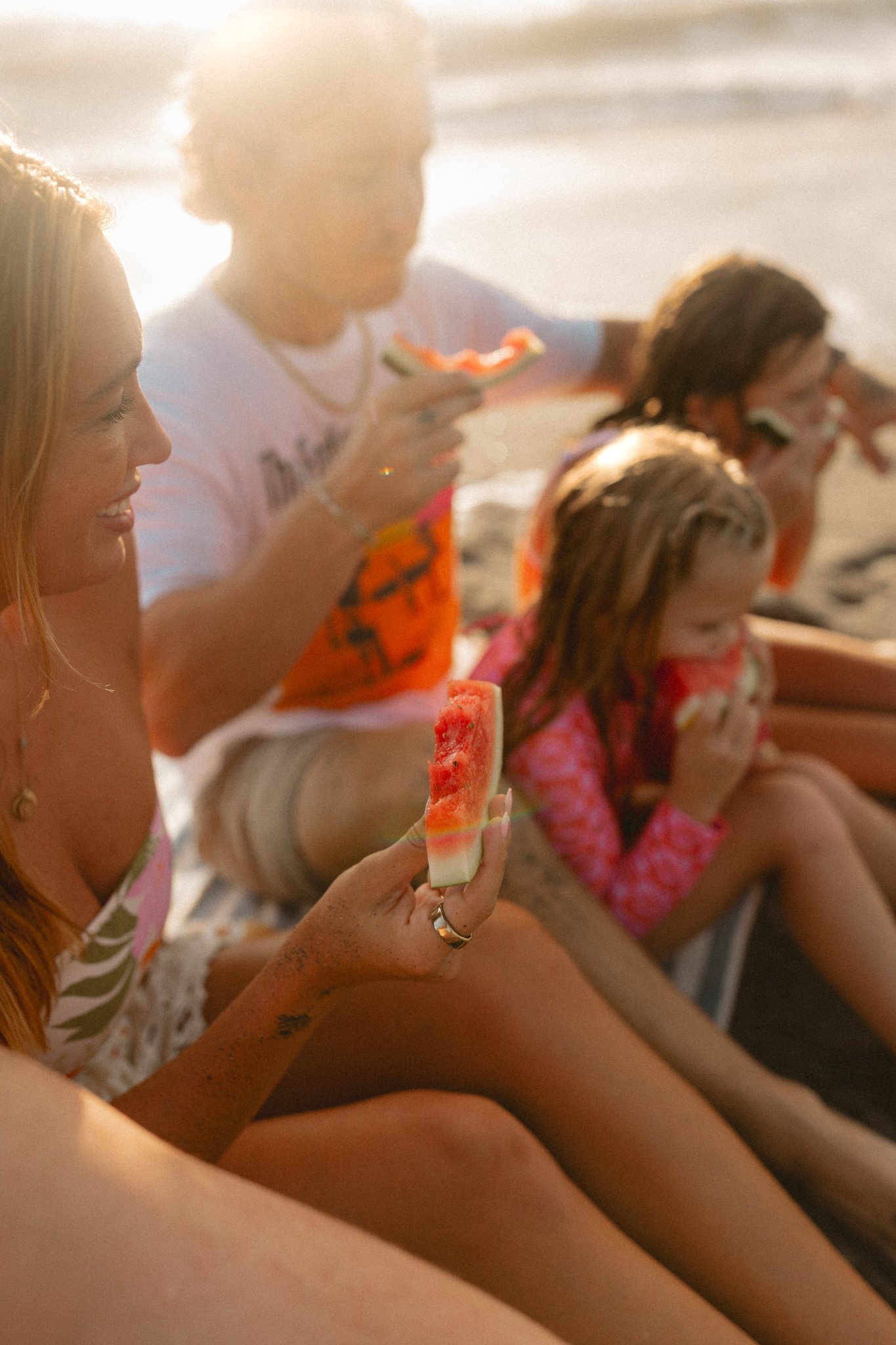Four people, two women and two children, sitting on the beach eating watermelon slices in the late afternoon sunlight.
