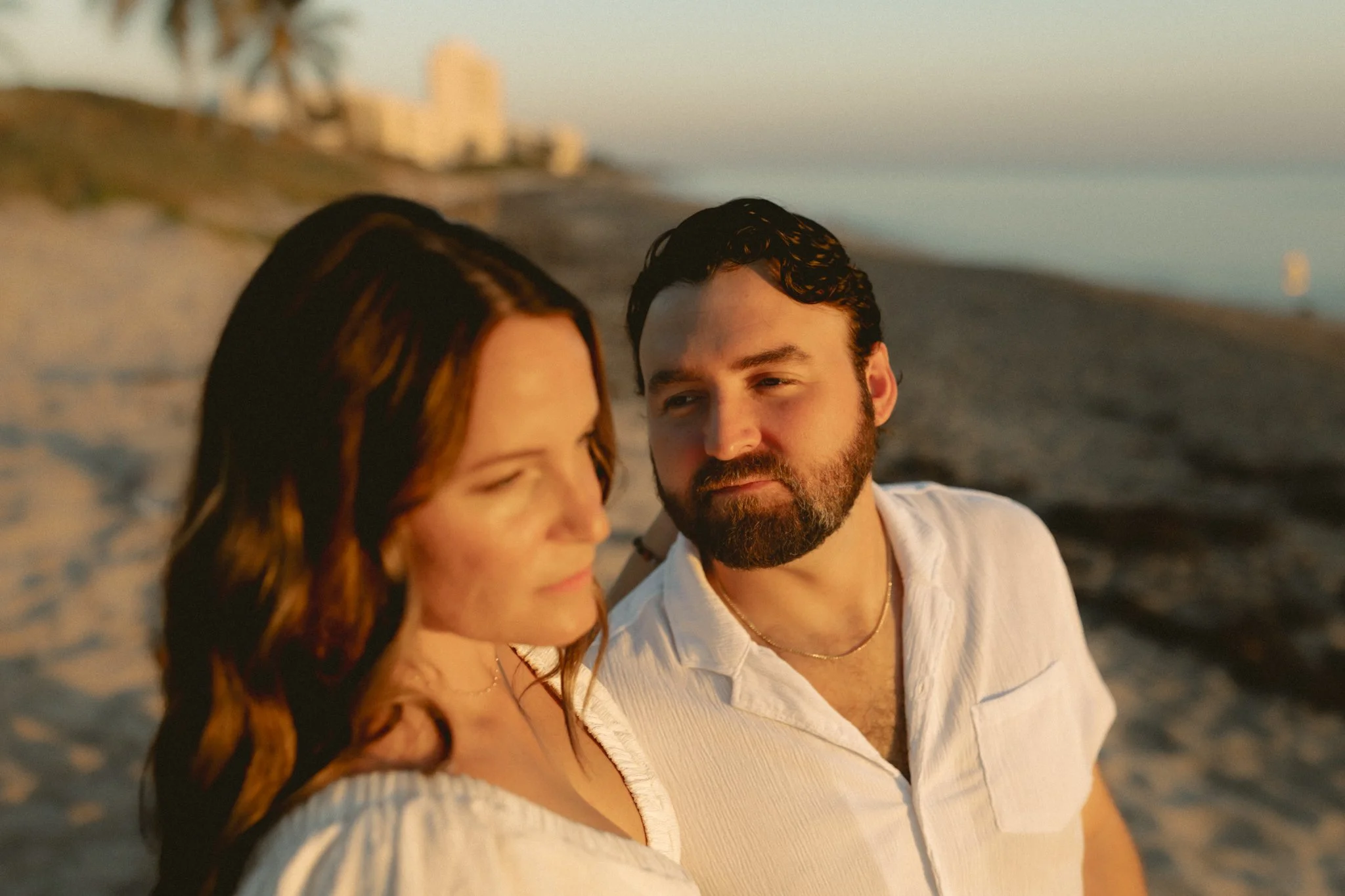A man and woman standing close together on a beach at sunset, with the city skyline visible in the background.