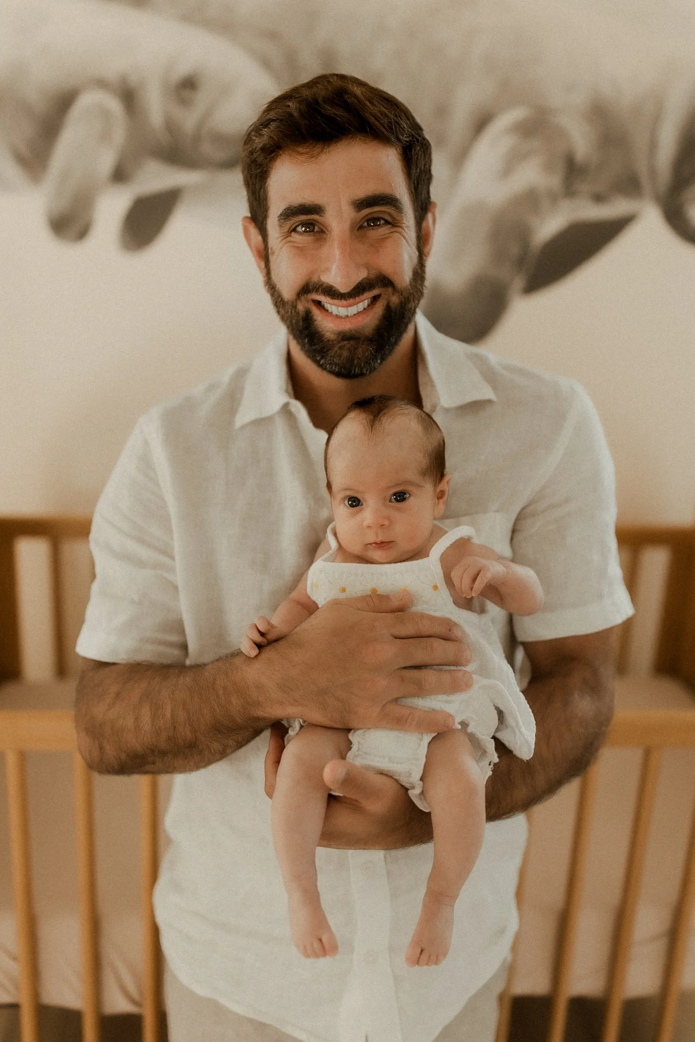 Smiling man holding a baby in a nursery with a wooden crib in the background.